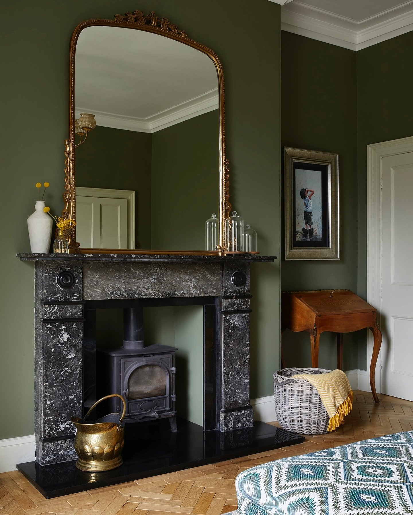 A classic black and white veined marble fireplace surround in a room with deep olive green walls, featuring a large gold-framed mirror and a wood-burning stove inside.