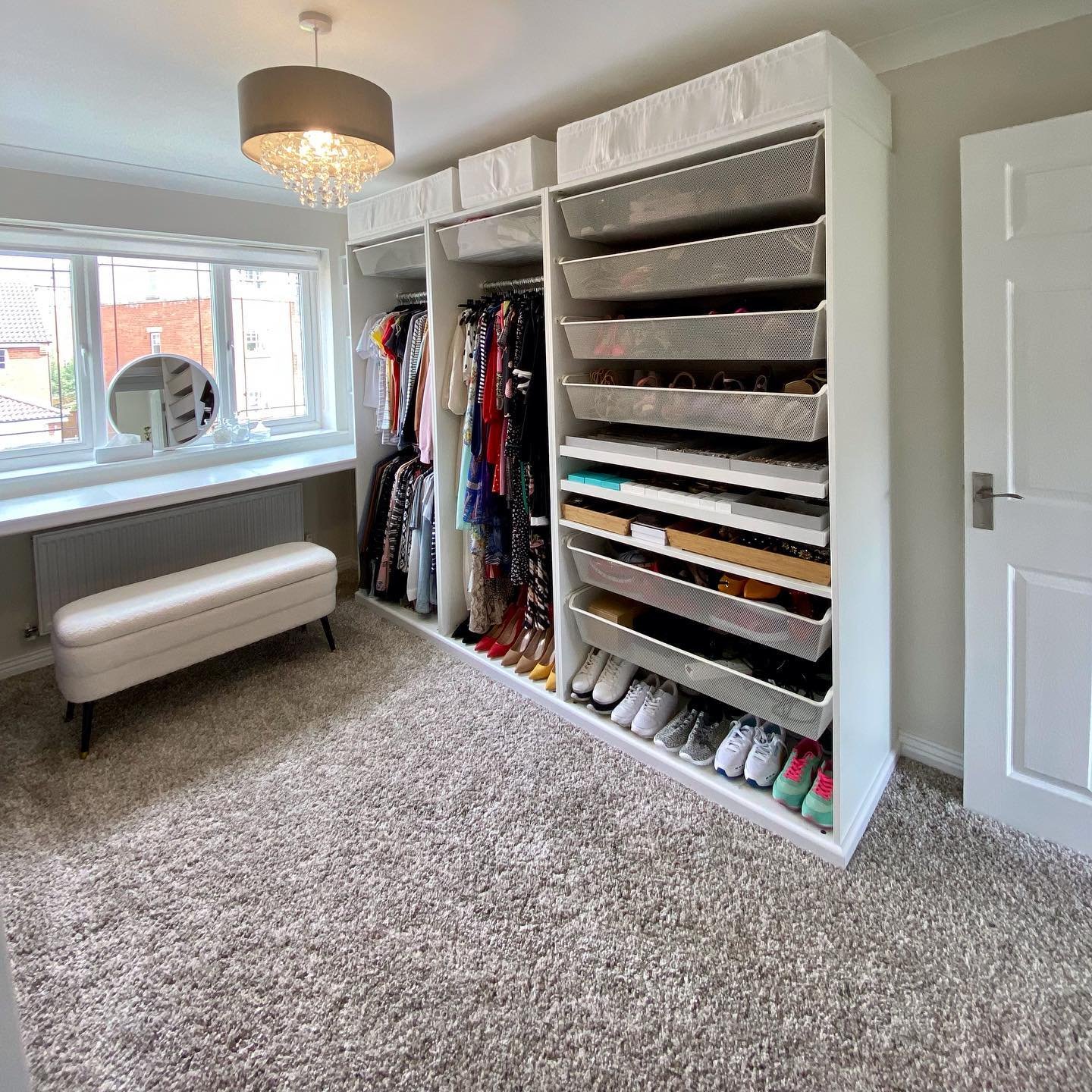 A functional, well-lit dressing room featuring custom white storage units with open shelving, mesh pull-out drawers for shoes, and a bench by the window.
