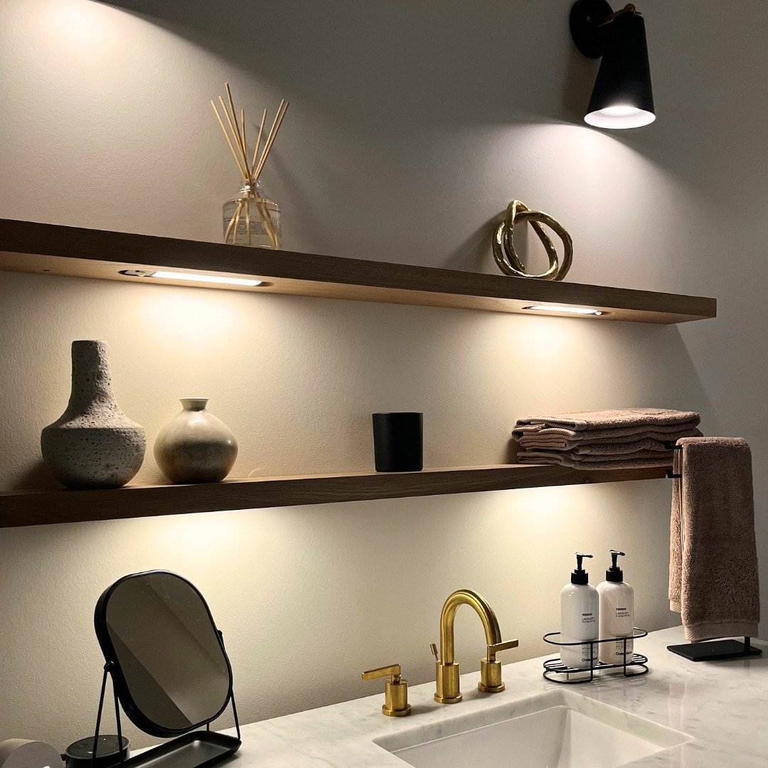 Long, thin wood floating shelves with under-mount strip lighting creating a warm glow above a bathroom sink.