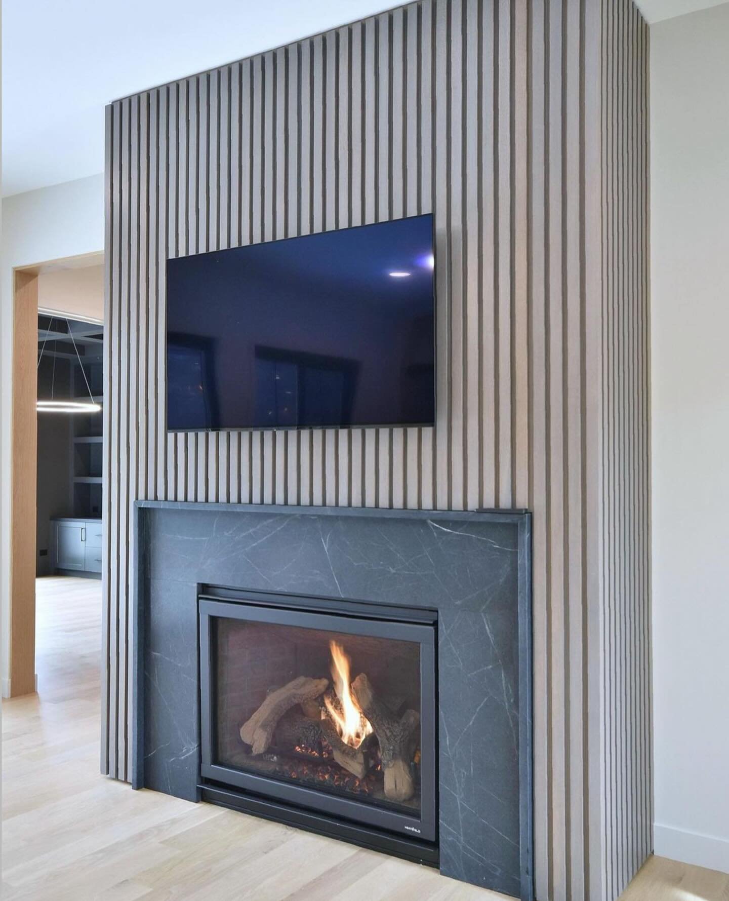 Freestanding fireplace accent wall clad in thin, vertical light wood slats with a recessed television and a dark soapstone fireplace surround.