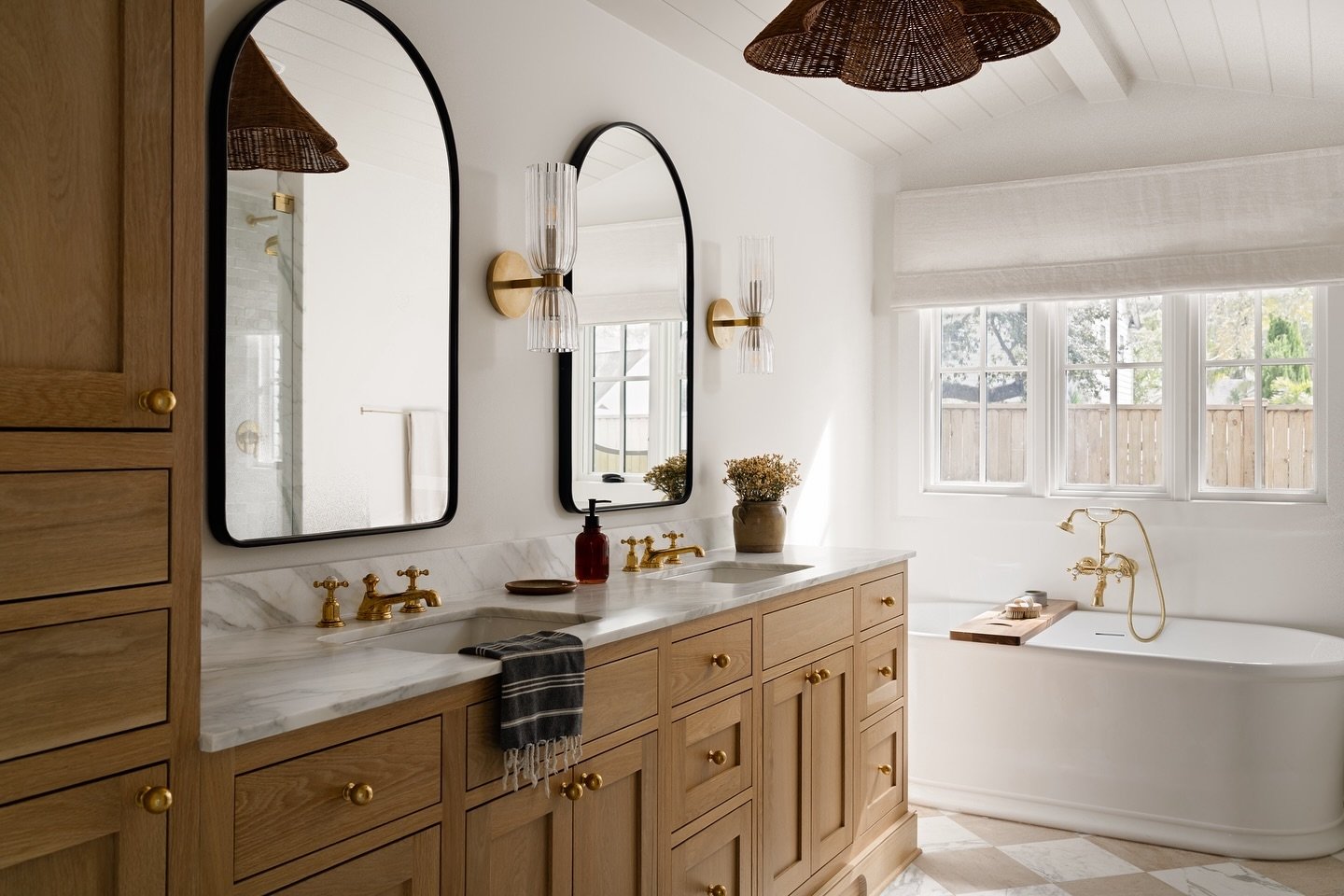 Double-sconces with brass bases and clear fluted glass shades flank two black-framed mirrors over a wood vanity with gold faucets and marble countertop.