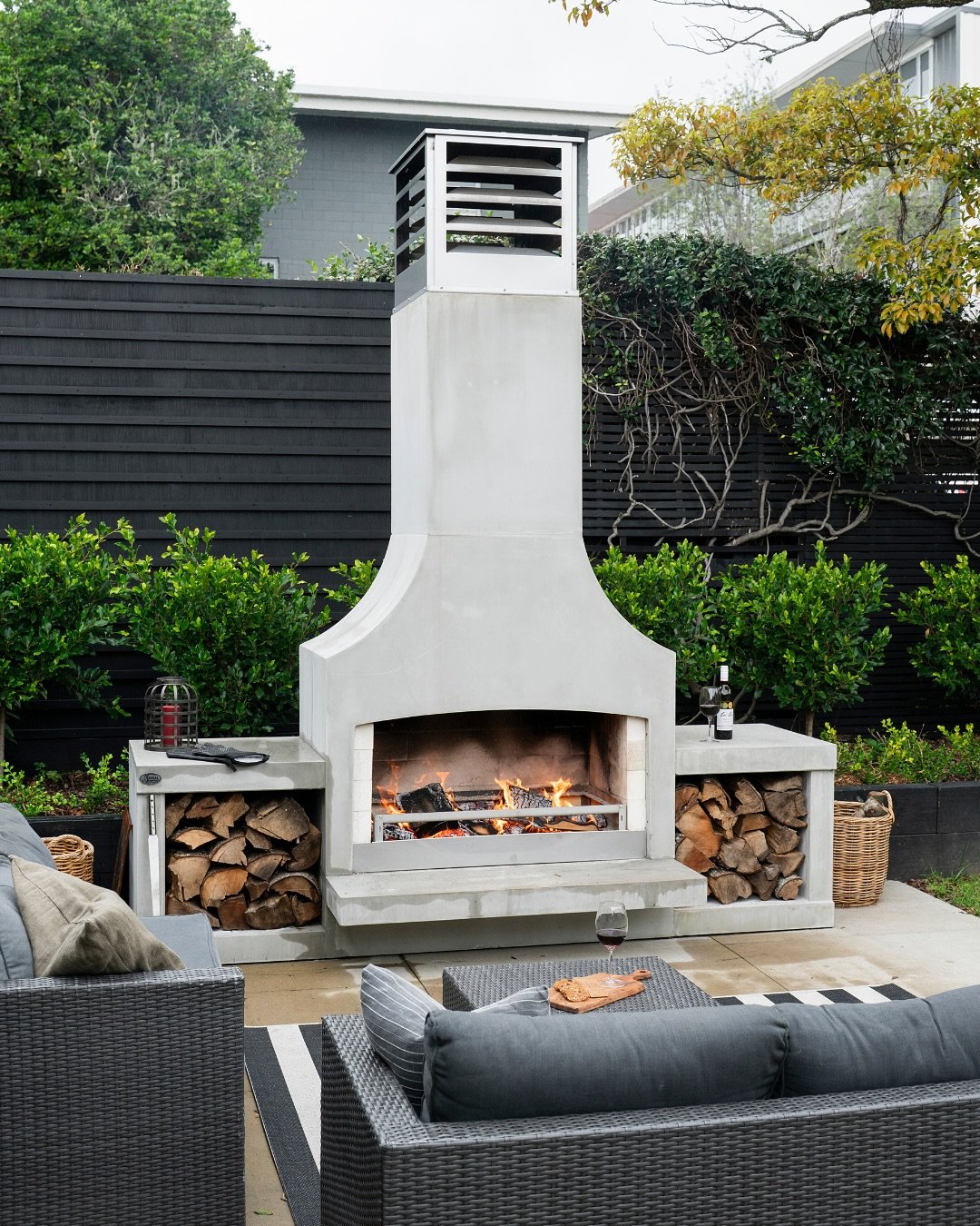A tall, modern pre-fabricated concrete outdoor fireplace with a flared base and integrated log storage wings.