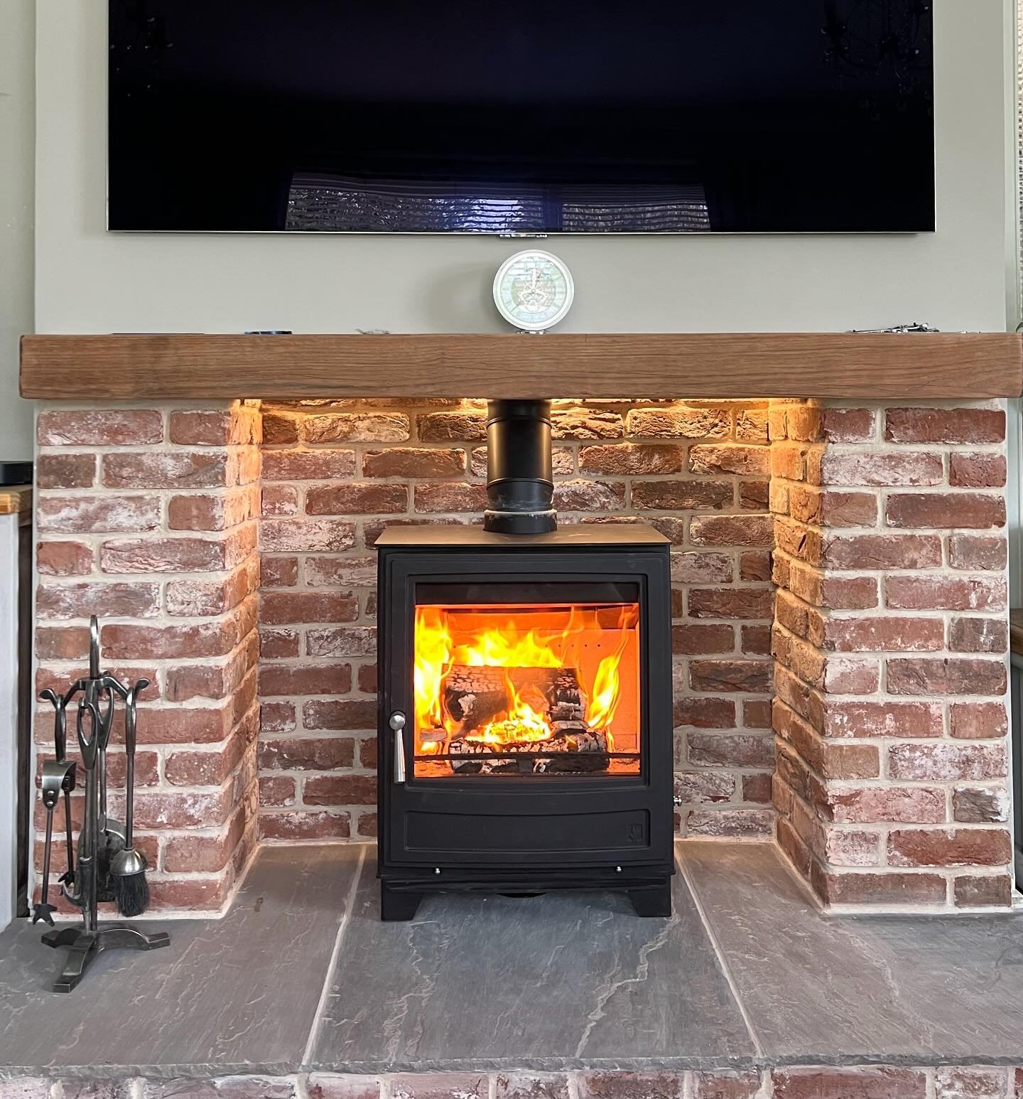 A rustic brick fireplace surround featuring natural red tones and a floating wood mantel. A black wood stove sits on a gray slate stone hearth.