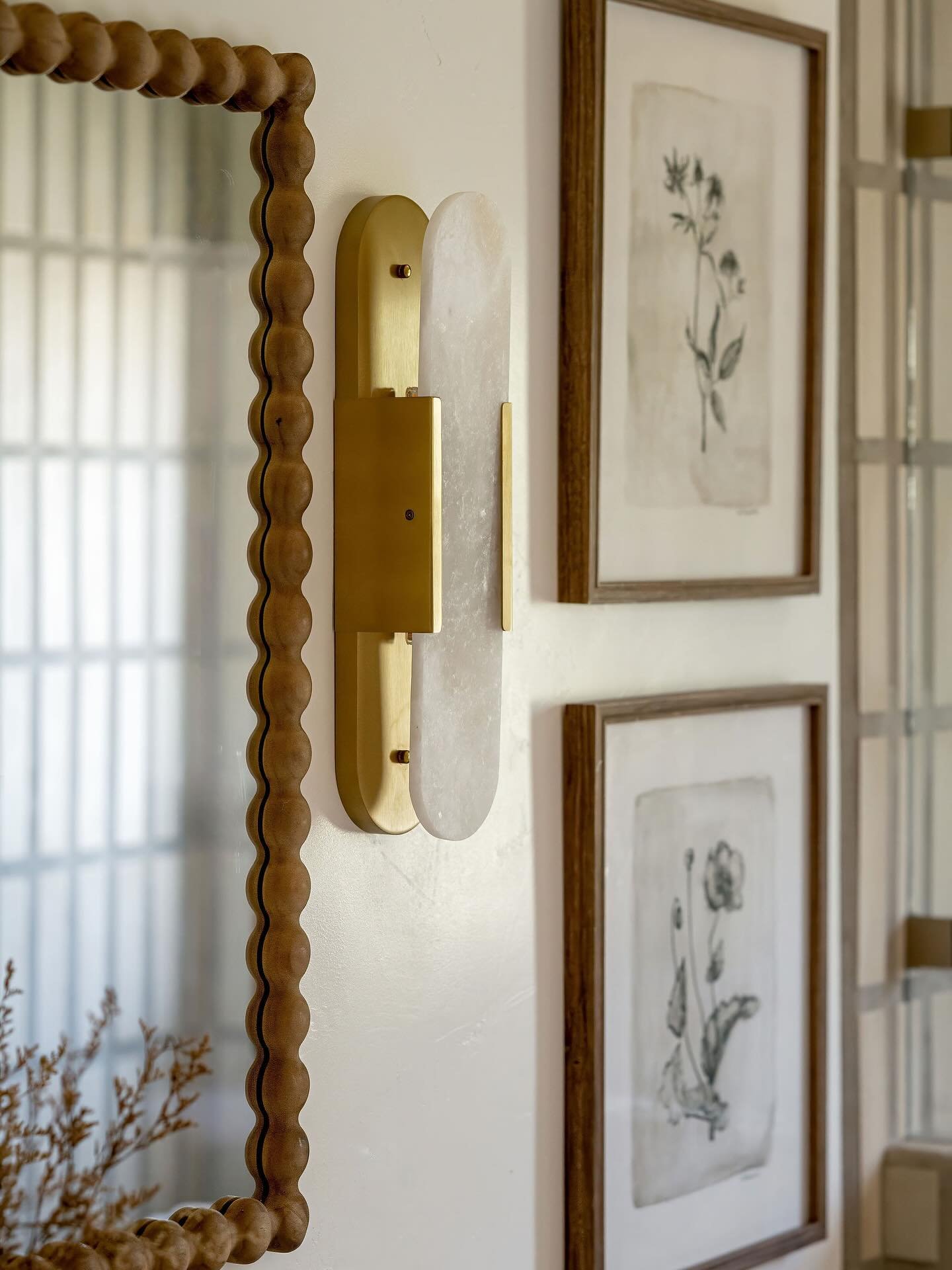 Vertical wall sconce with a long, thin slab of white alabaster stone mounted on a rectangular brass backplate, next to a wood beaded mirror.