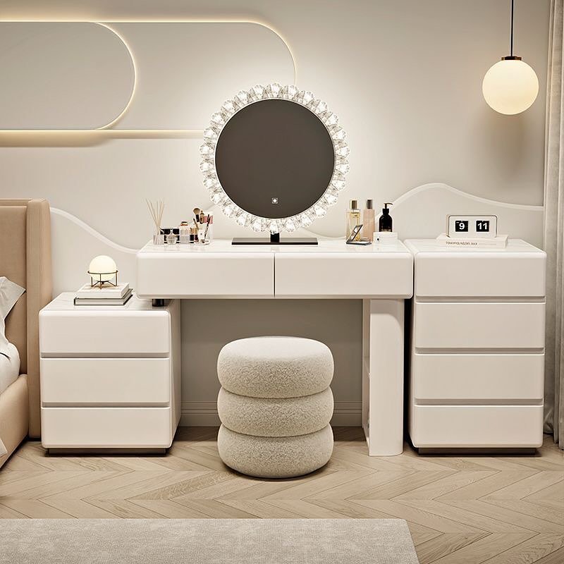 A modern white dressing table setup composed of a central desk and two large side storage chests, featuring a circular, crystal-rimmed, backlit mirror and a stacked white bouclé ottoman.
