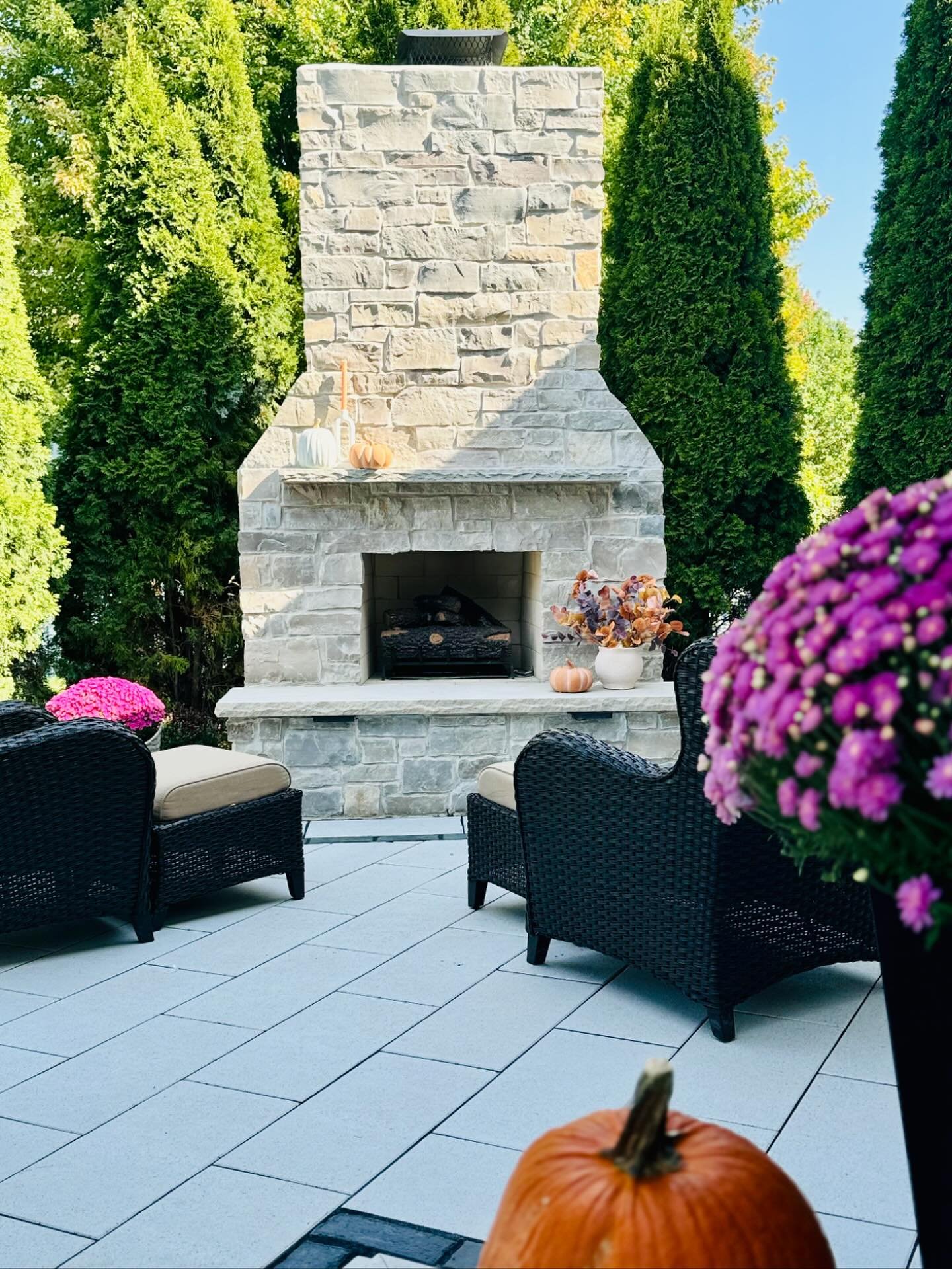 A tall, traditional outdoor fireplace built with light gray fieldstone on a white paver patio, surrounded by tall greenery.