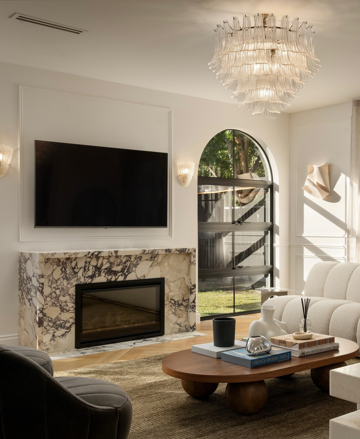 A modern living room with a thick, floating marble mantle featuring dark purple and brown veining, centered below a television and near a large arched window.