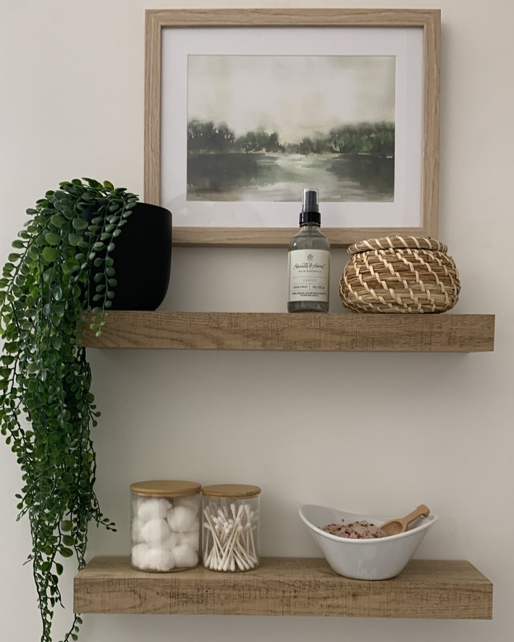 Two rustic light-wood floating shelves on a white wall, holding plants, art, and glass jars.