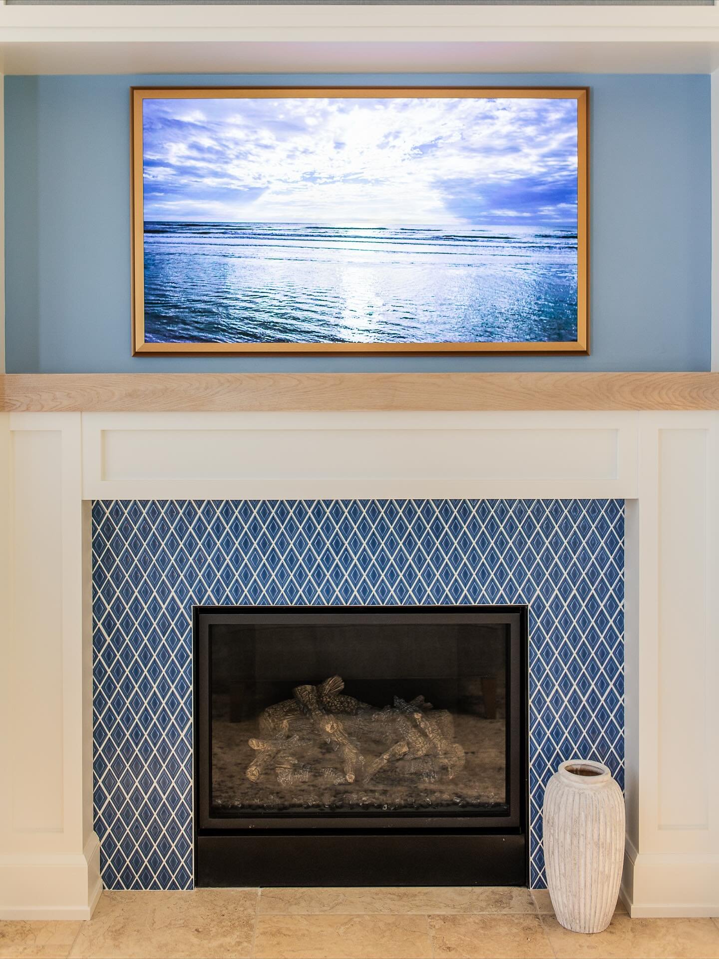 A coastal-style fireplace with a white surround and natural wood mantel, featuring navy blue diamond tiles with a contrasting white geometric pattern.