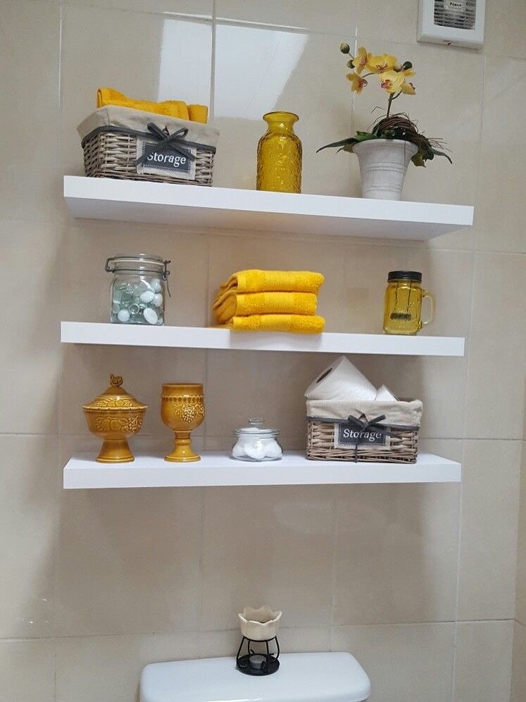 Three long white floating shelves stacked on a tiled wall, decorated with yellow towels and baskets.