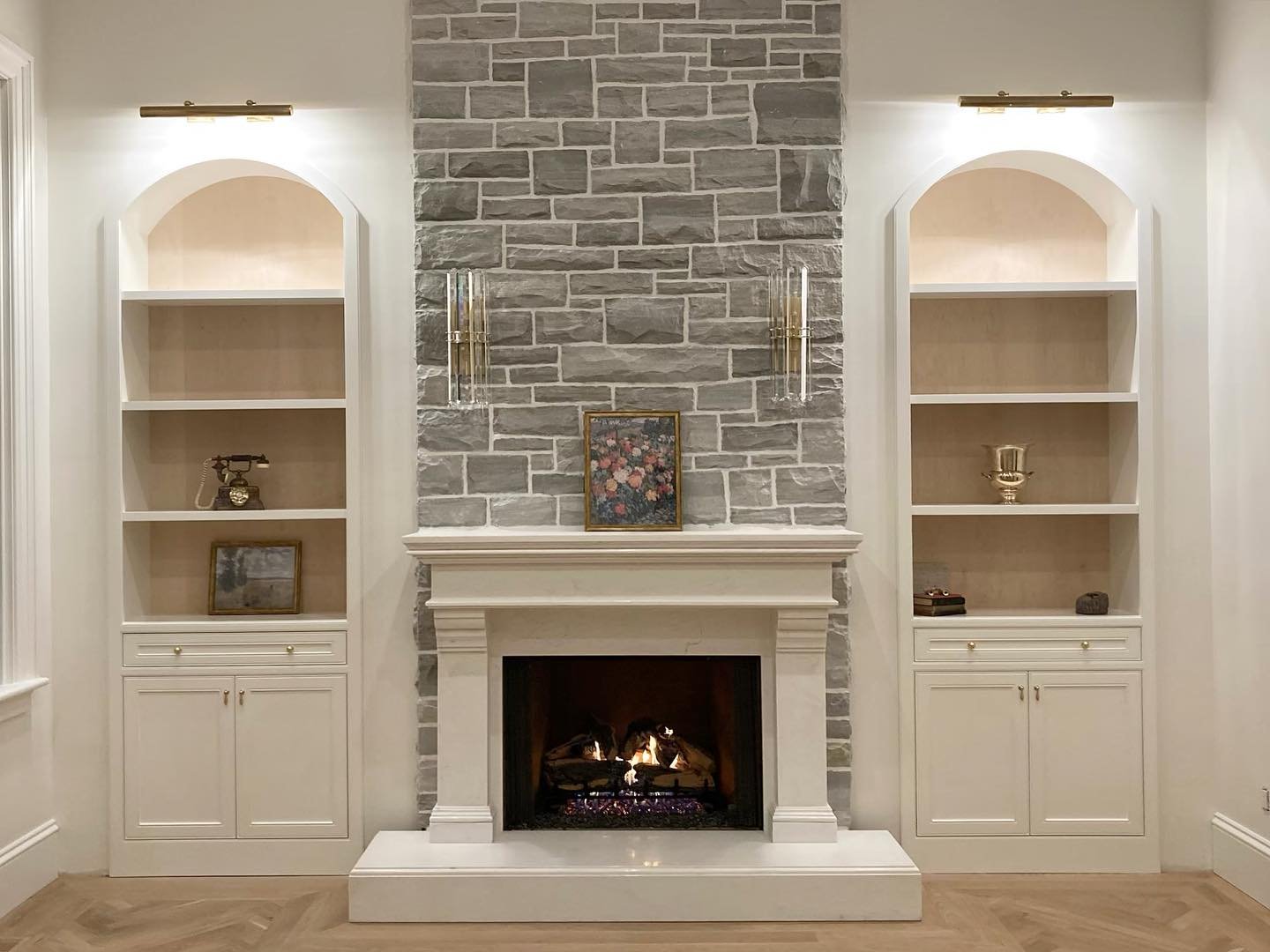 White arched built-in shelves with brass lighting flanking a grey stone fireplace.