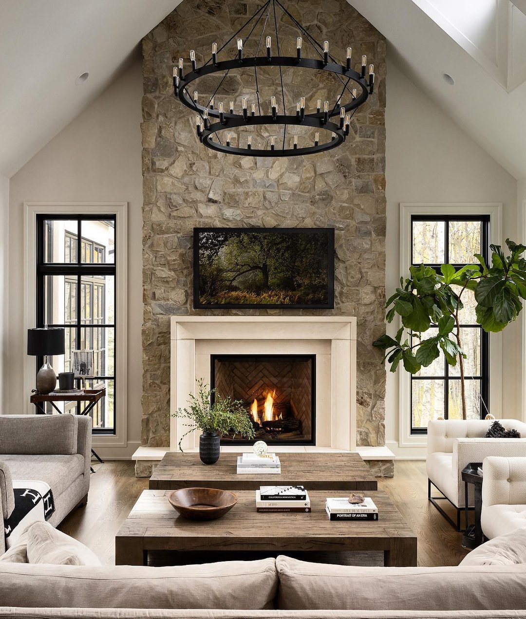 A tall, vaulted-ceiling living room with a prominent fireplace constructed from rough, grey fieldstone, featuring a light, smooth stone surround, a herringbone firebox interior, and a large black chandelier.