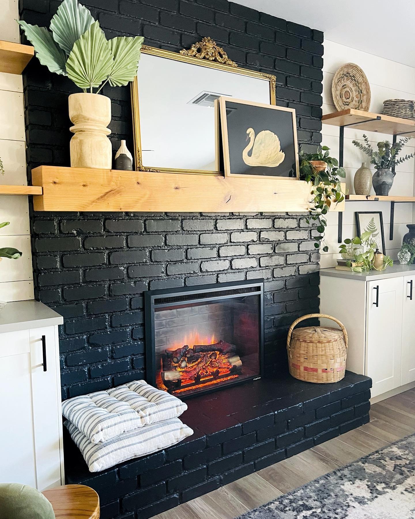 A brick fireplace painted deep matte black with a light wood floating mantel. White cabinets and shelving flank the dark brick structure.