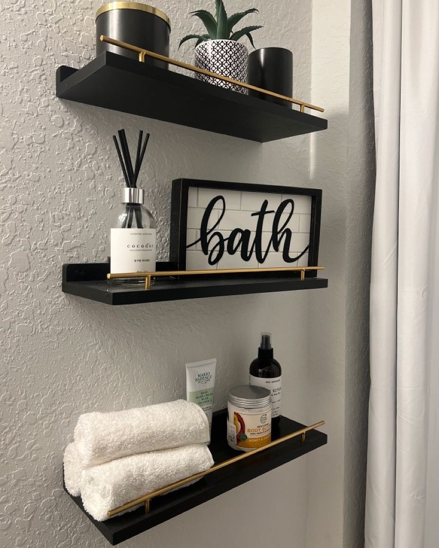 Three black floating ledge-style shelves with gold gallery rails mounted on a textured grey wall.