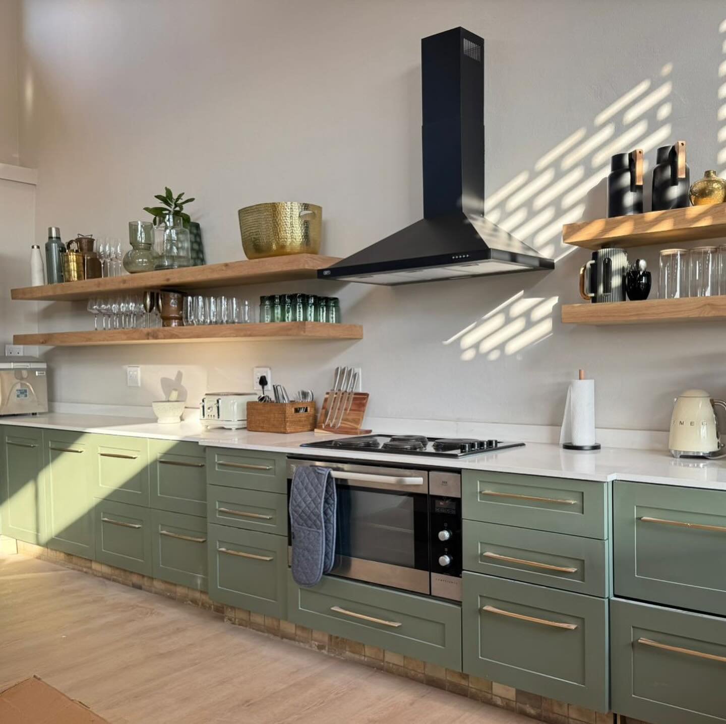 Light wood floating shelves on either side of a black range hood in a kitchen with sage green cabinets.