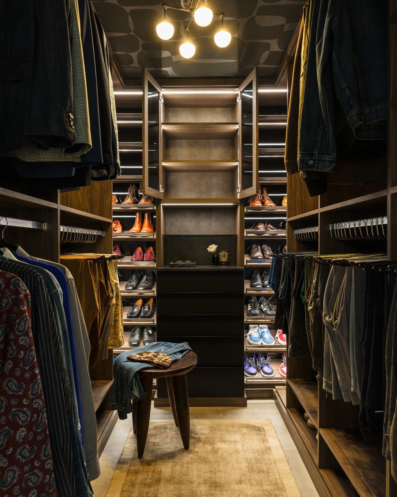 A masculine walk-in closet featuring dark wood cabinets, a black reeded central drawer unit, lighted shelves for shoes, and a unique patterned ceiling.