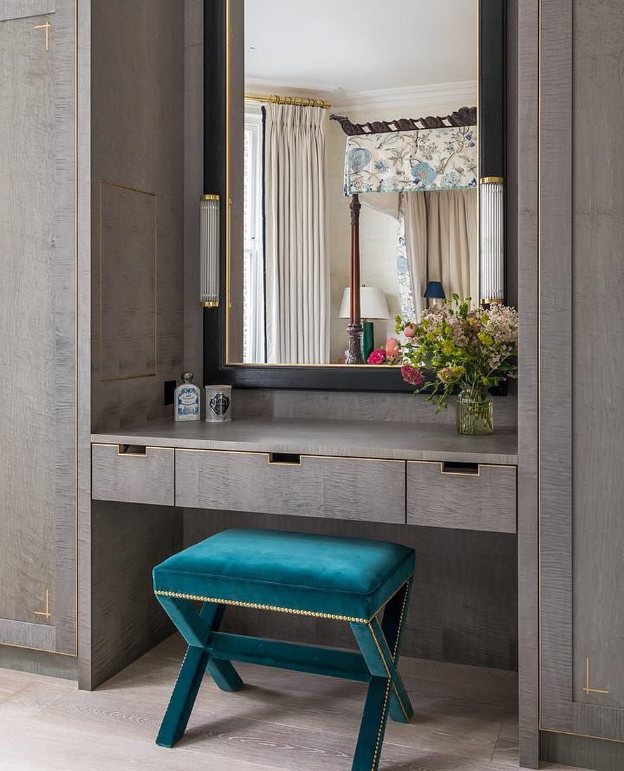 A built-in gray wood dressing table with three drawers and a tall mirror flanked by vertical sconces, perfectly fitted into wood-paneled walls, with a striking teal velvet X-leg stool.