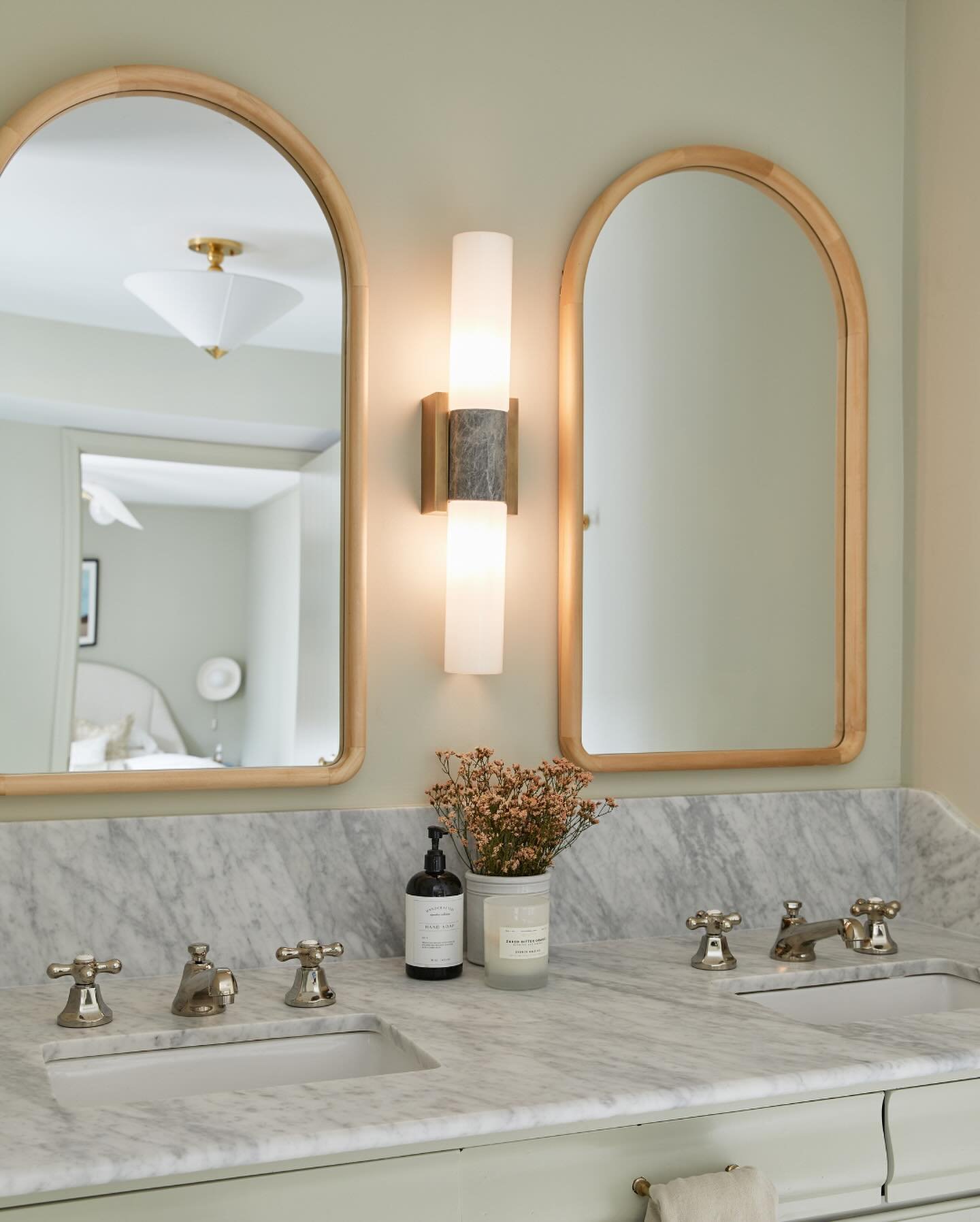 Vertical double-cylinder sconce featuring frosted glass and a dark stone or marble band, centered between two light wood-framed arched mirrors.