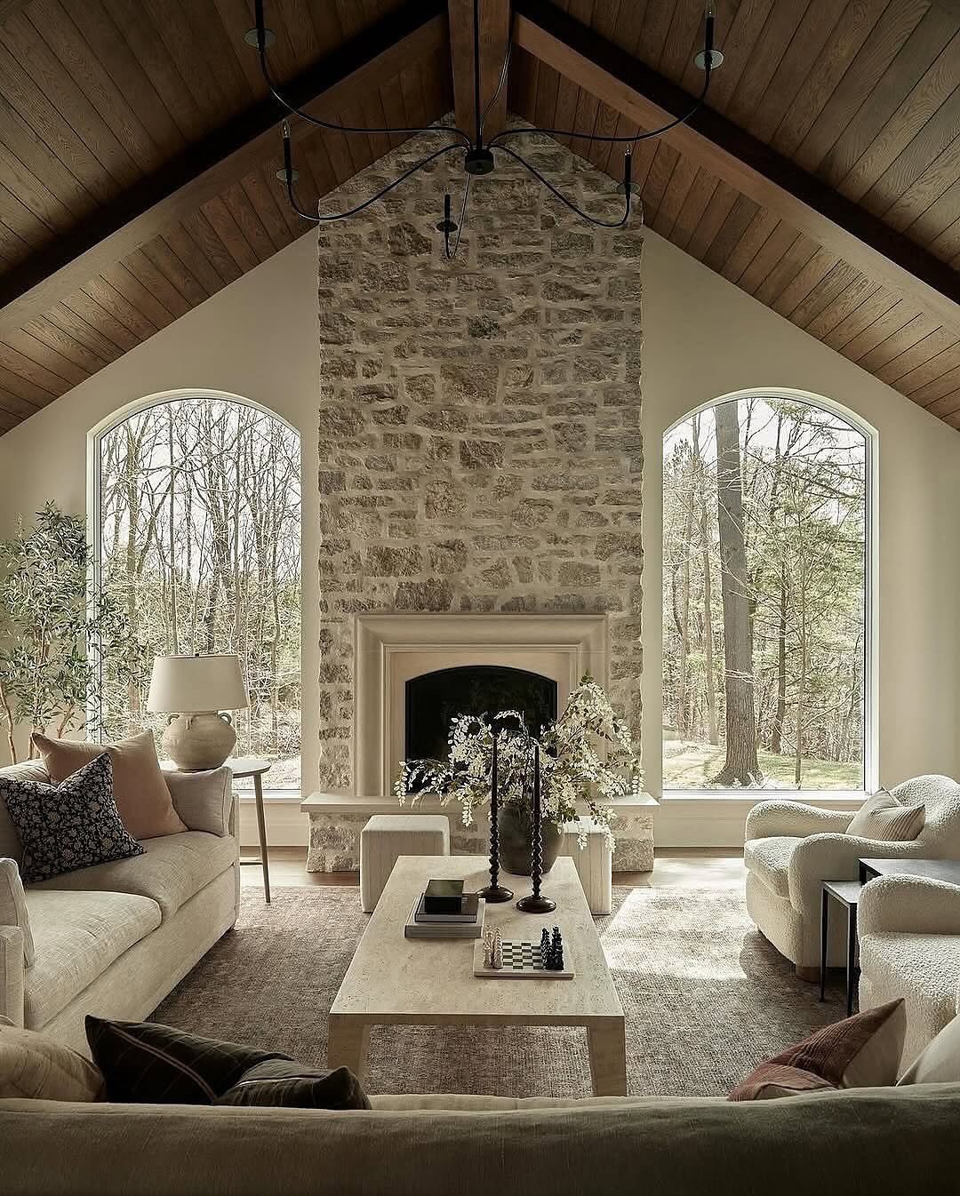 A tall, vaulted-ceiling living room with a stone fireplace made of rough, pale fieldstone, featuring a classic, ornate, smooth light-colored stone surround, flanked by two large arched windows.
