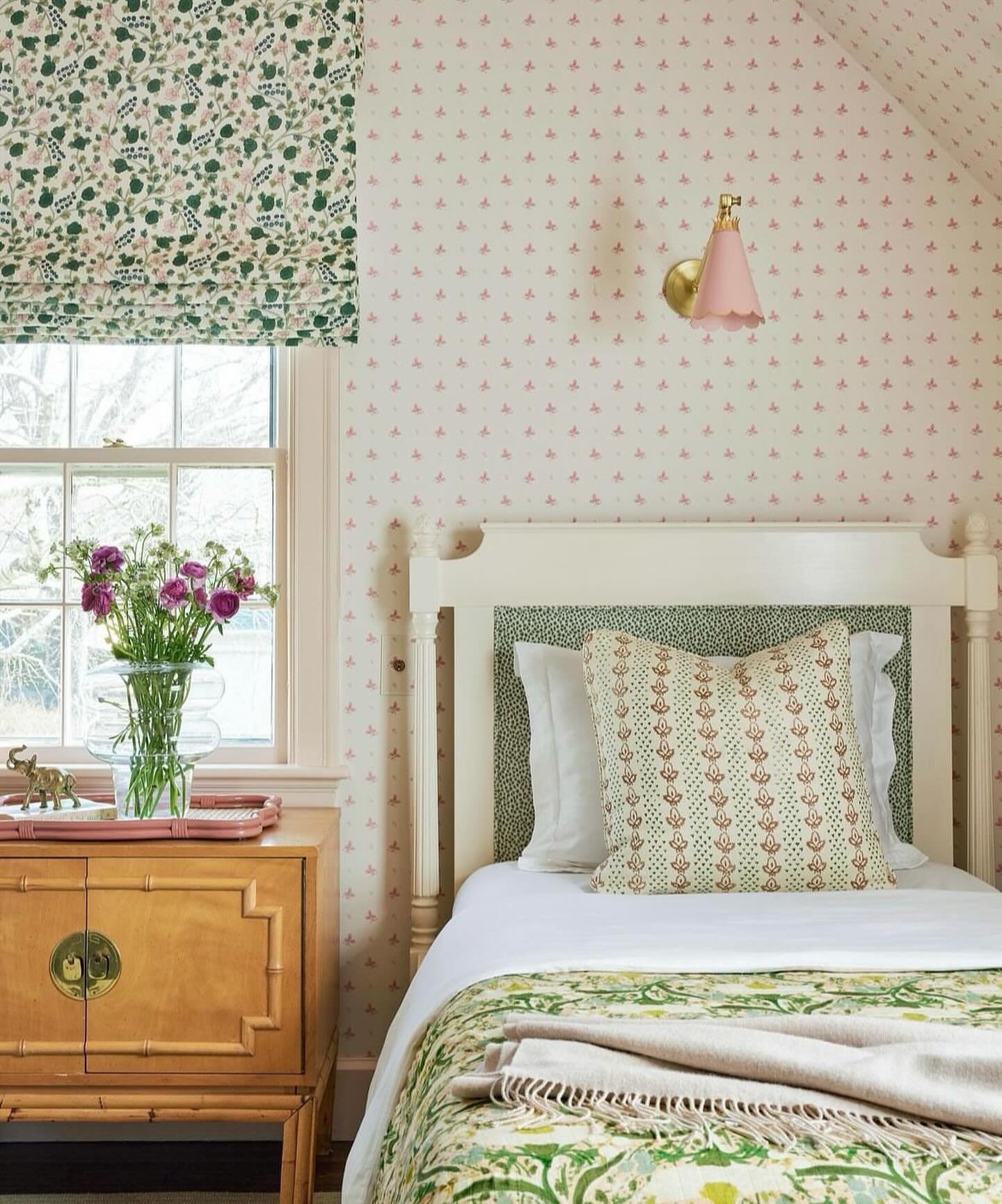 A cozy single bedroom featuring a subtle pink and cream patterned wallpaper, a green and white botanical Roman blind, and a Chinoiserie-style nightstand.
