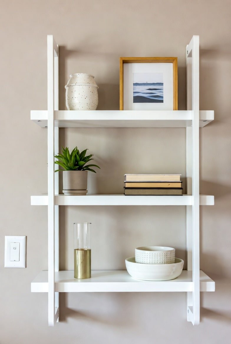Three-tier white ladder style wall shelf unit on beige wall with decor