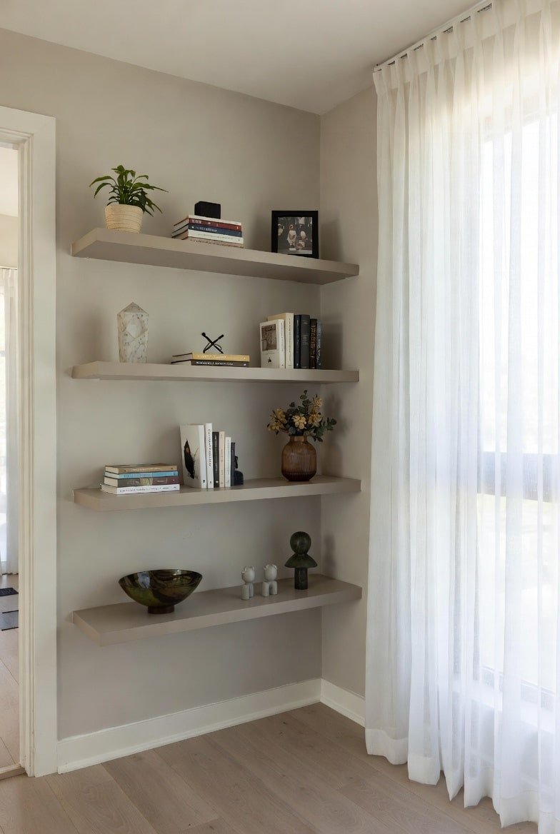 Four greige floating shelves installed in a wall niche next to white sheer curtains.
