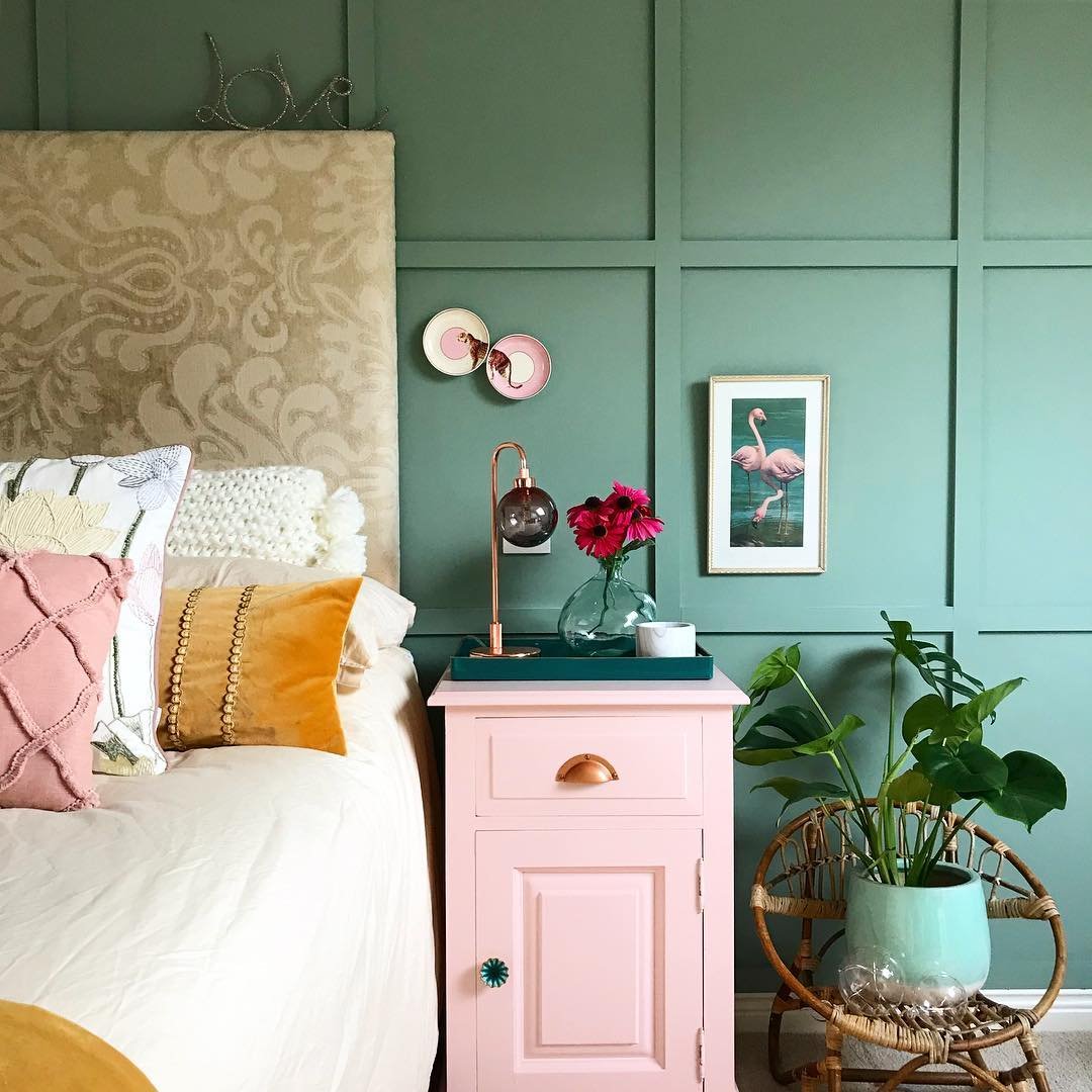 A bedroom close-up featuring a paneled wall painted in a cheerful sage green, a bright pink nightstand, and a large upholstered headboard with a subtle pattern.