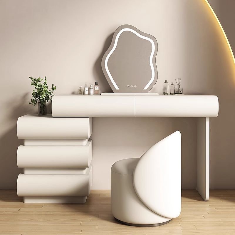 An ultra-modern white dressing table with a rounded design, a separate four-drawer chest with a stacked cylindrical shape, and an irregular, wavy backlit mirror, paired with a curved white leather stool.