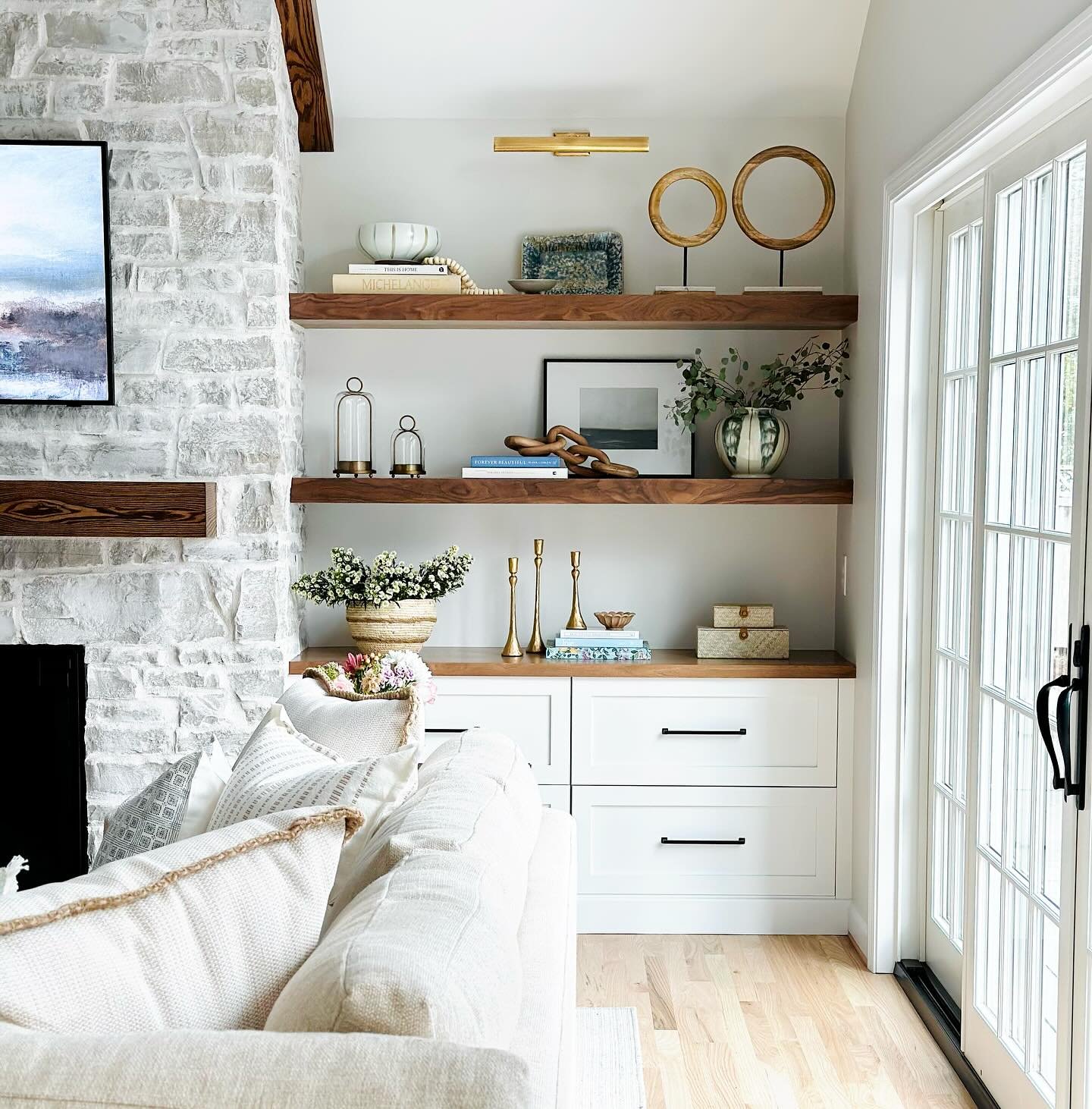 Thick walnut floating shelves and white drawers next to stone fireplace with brass accents