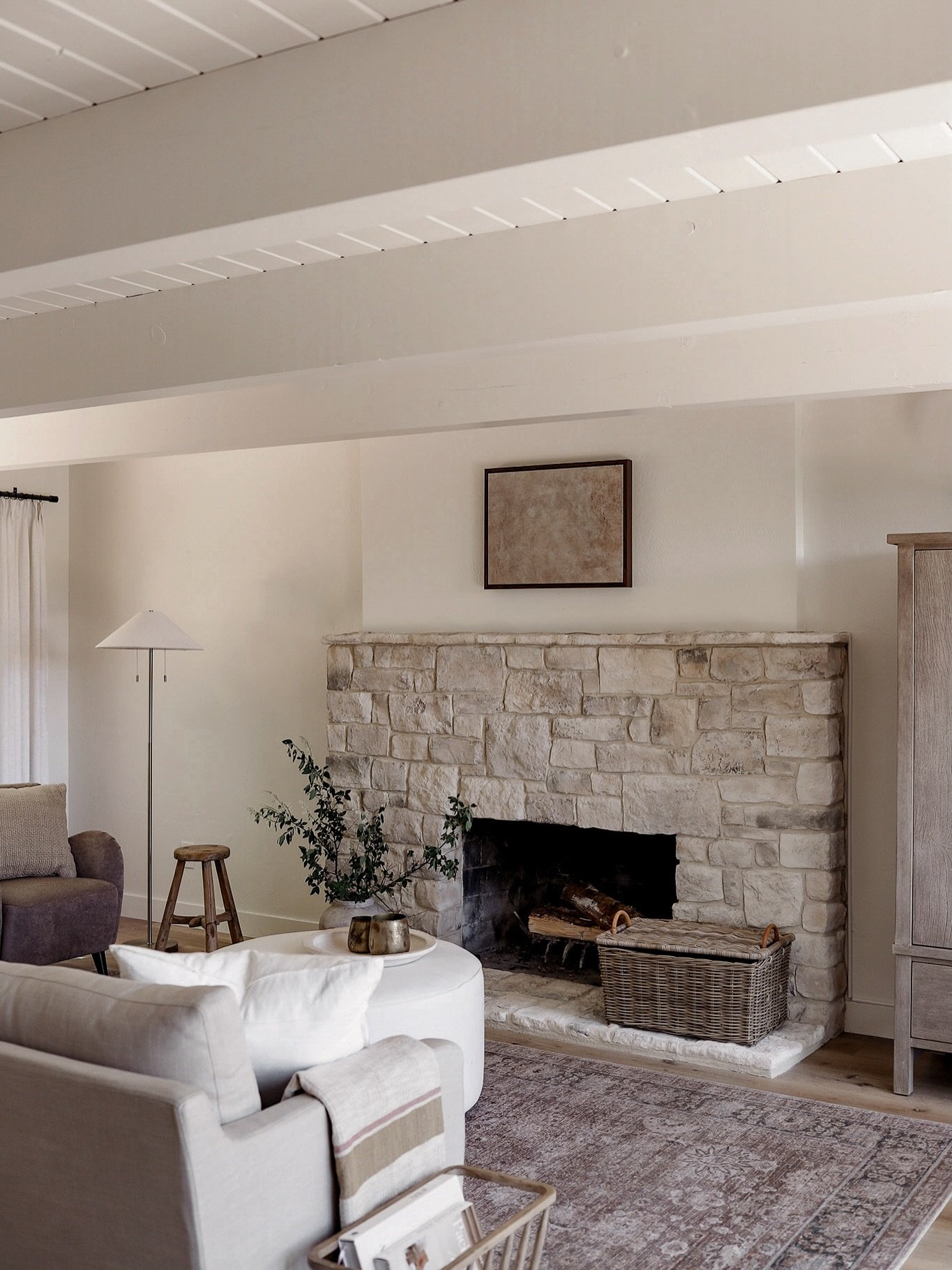 A wide, low stone fireplace made of rough, rectangular pale limestone blocks, featuring a flush firebox, a thick stone hearth, and a wicker log basket beside it in a relaxed living room.