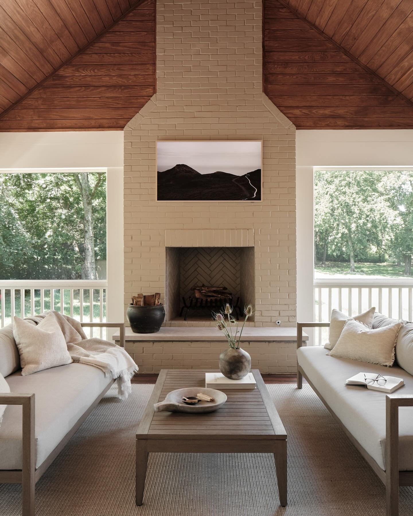 Traditional style fireplace made of brick painted a light taupe color, set beneath a dark wood vaulted ceiling on a covered porch.