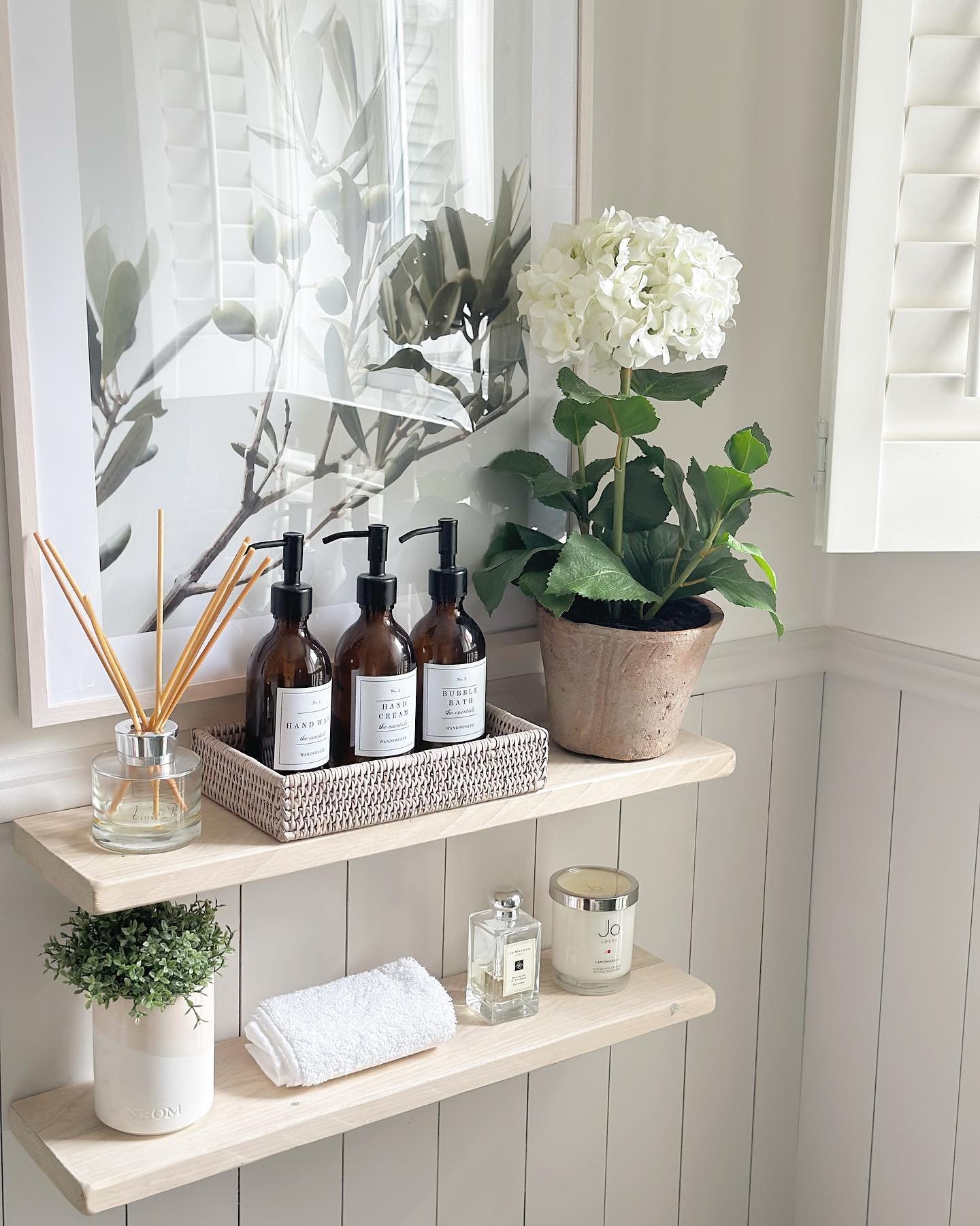 Two light wood floating shelves mounted on white wainscoting, holding spa-like items including soap bottles, plants, and candles.