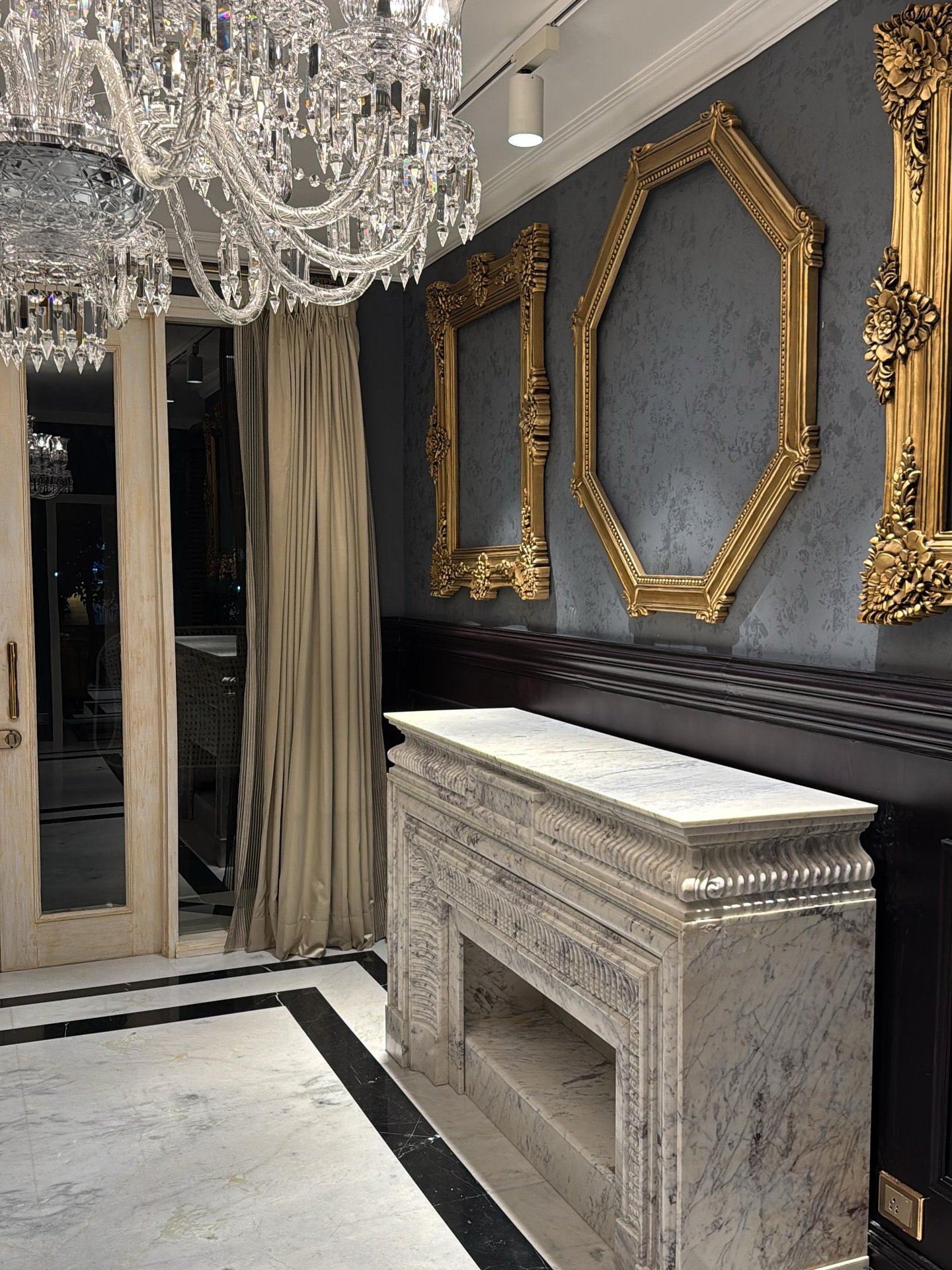 An entry hall featuring a highly carved white marble fireplace mantel used as a console, set against a dark wall with ornate gold frames and beneath a crystal chandelier.