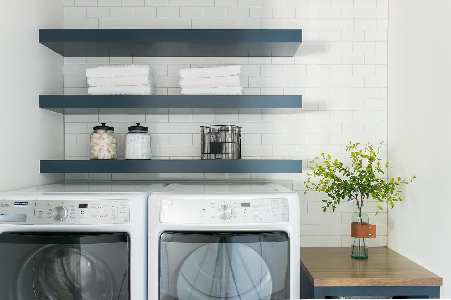 Dark blue floating laundry shelves mounted on white subway tile wall holding towels and glass jars
