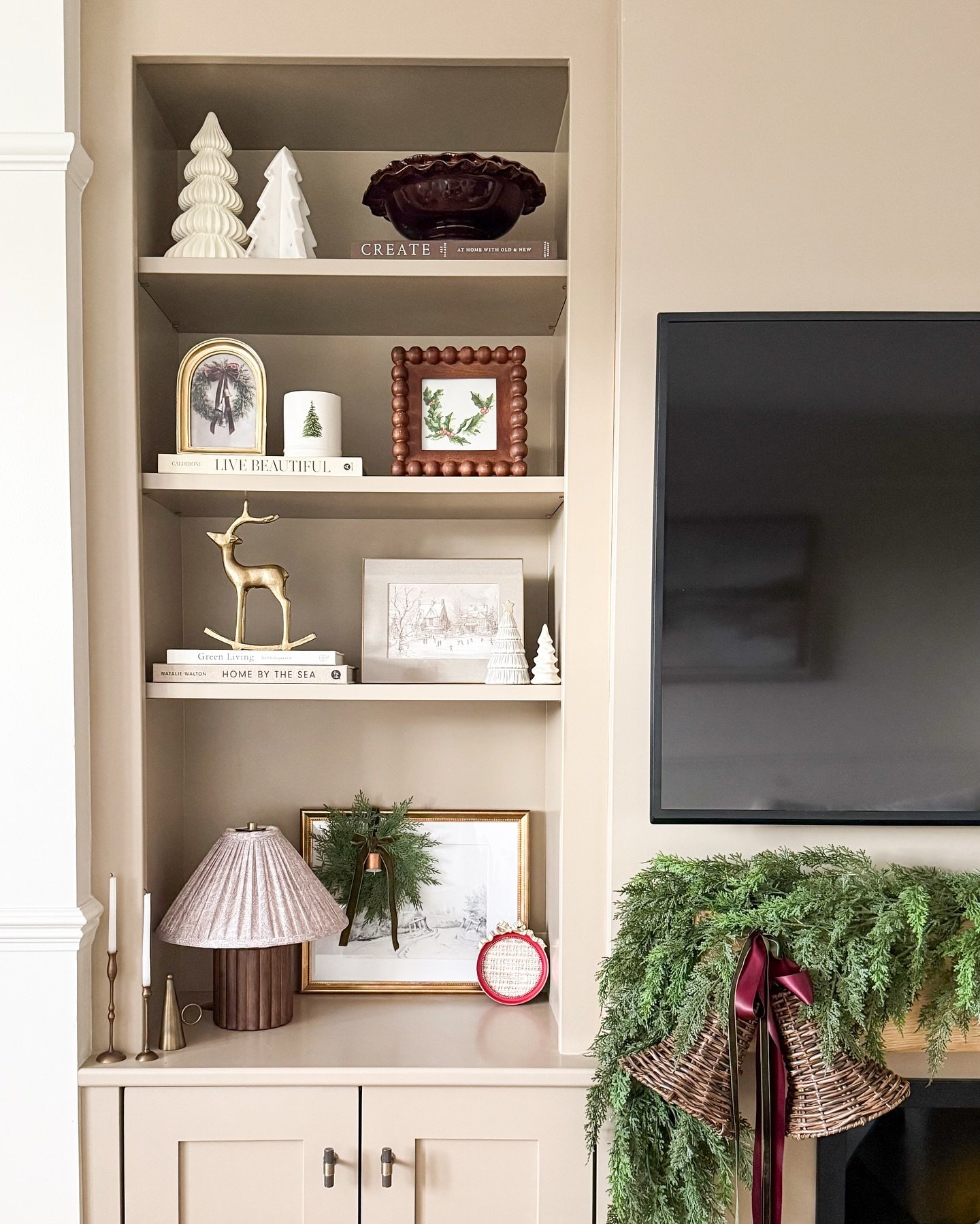 Beige built-in shelves decorated with white ceramic trees and festive art next to a TV.