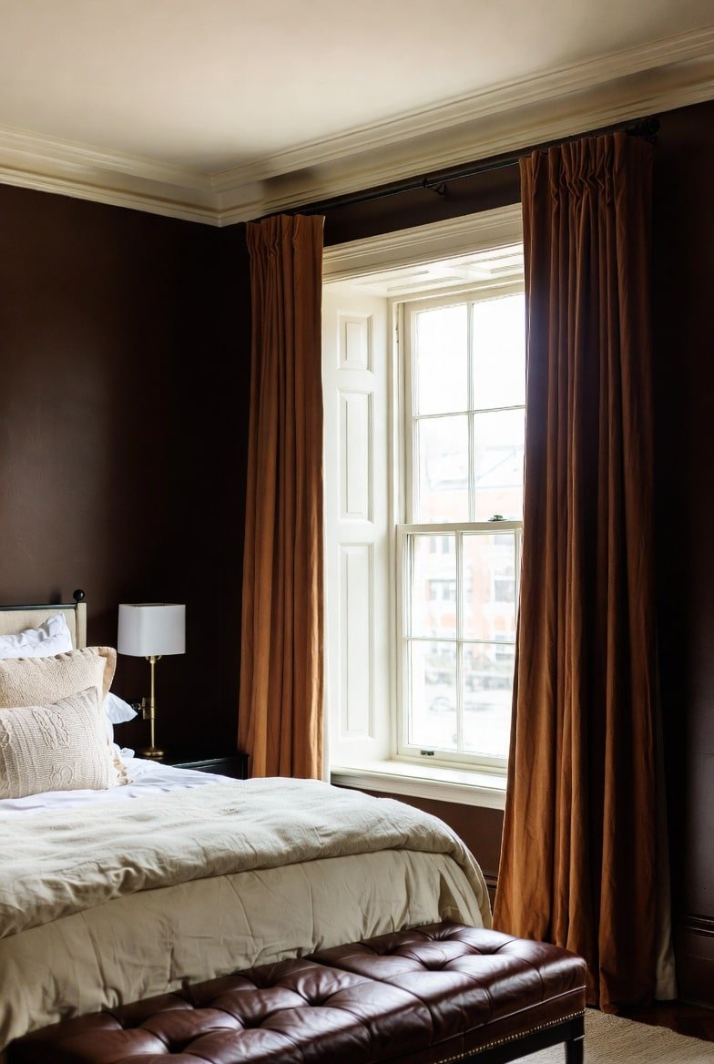 Dark brown bedroom with rust velvet curtains, white window shutters, and leather bench.