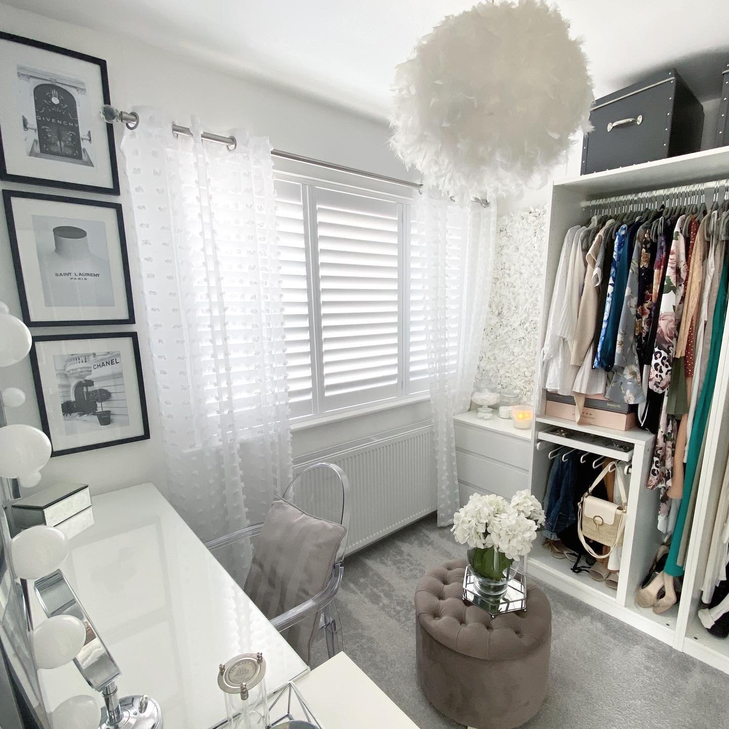 A small, bright dressing room and vanity area featuring an open white closet, a plush grey ottoman, a clear acrylic chair, and fashion-themed framed art.
