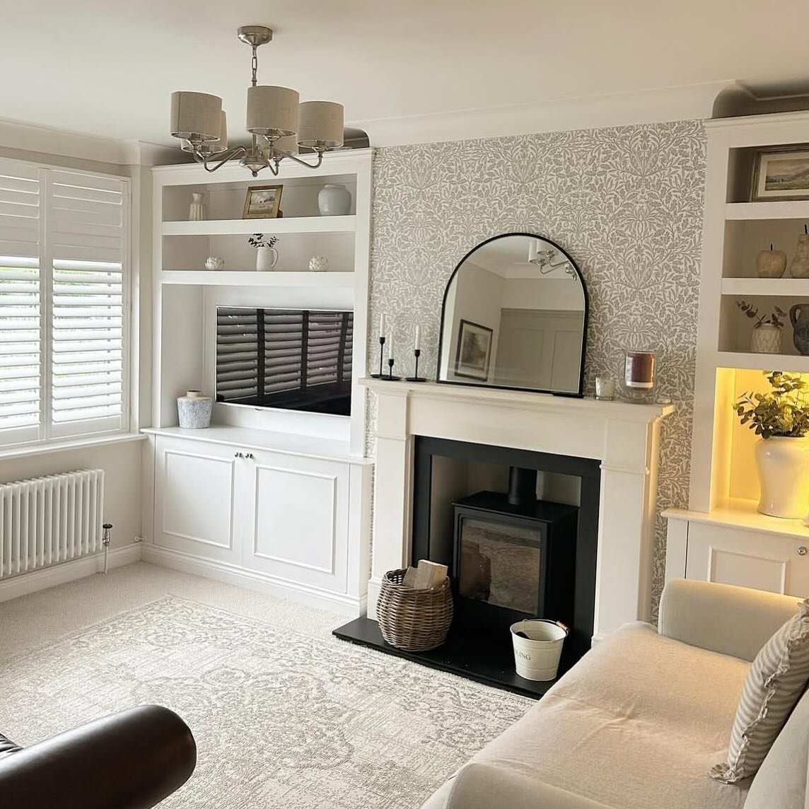 White built-in shelves flanking a fireplace with grey patterned wallpaper and an arched mirror.