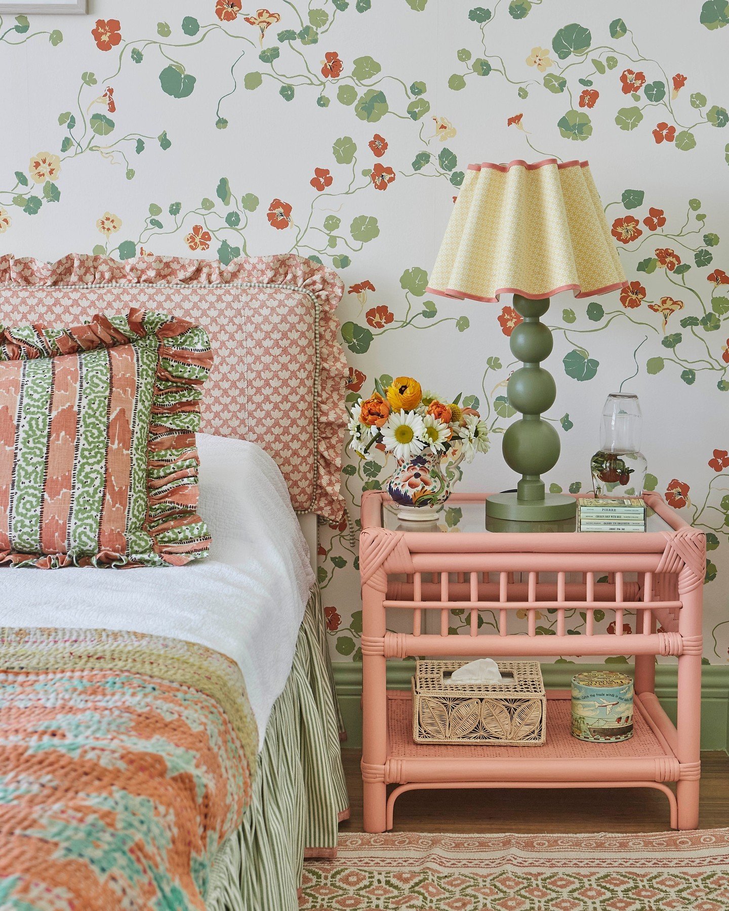A close-up of a bed and pink rattan nightstand against a wall covered in a botanical, vine-patterned wallpaper with green and coral flowers.