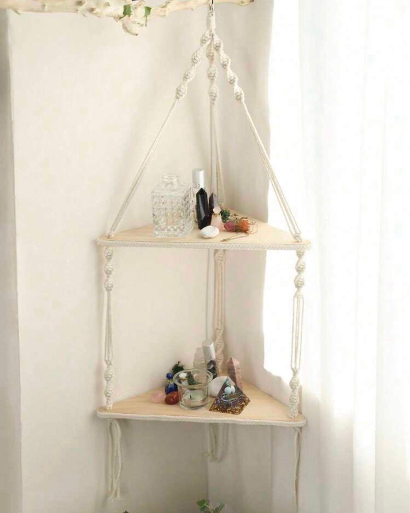 Two-tier light wood triangular shelf suspended by white knotted macramé rope in a corner.
