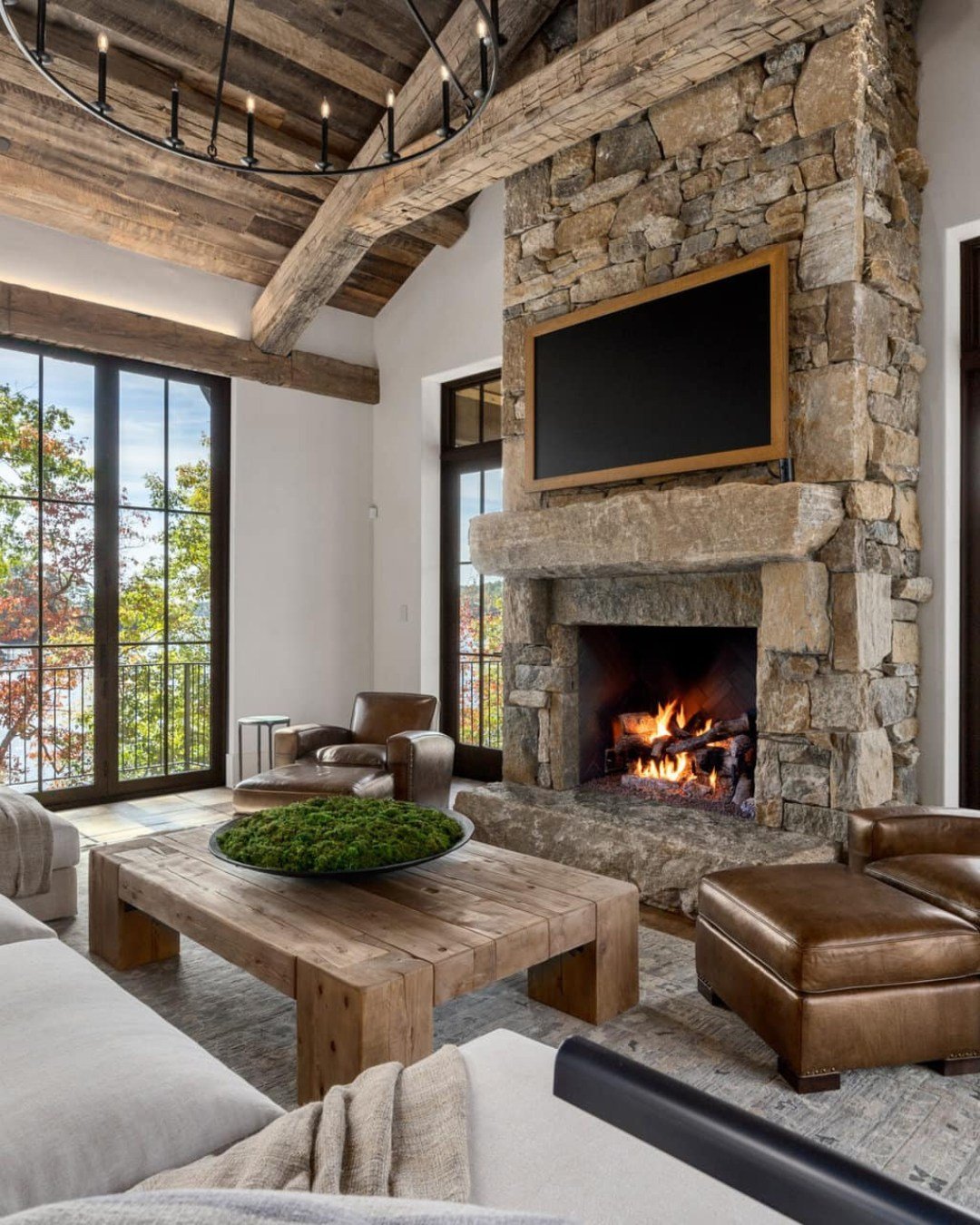 A massive, full-height fireplace constructed from chunky, rough-cut brown and grey fieldstone, featuring a large, protruding stone mantel and hearth, set in a room with vaulted timber ceilings and large windows.