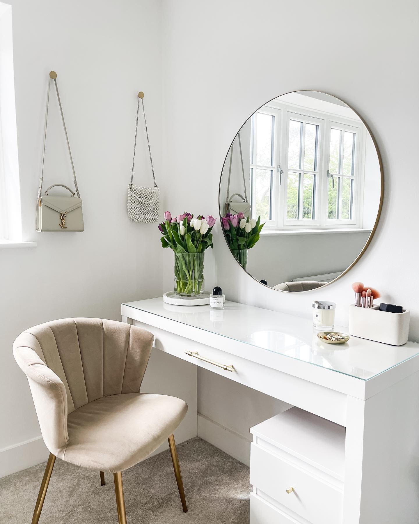 A bright, white high-gloss dressing table with a glass top, featuring a large gold-rimmed round mirror and a beige scalloped velvet chair with gold legs, in a minimalist room.