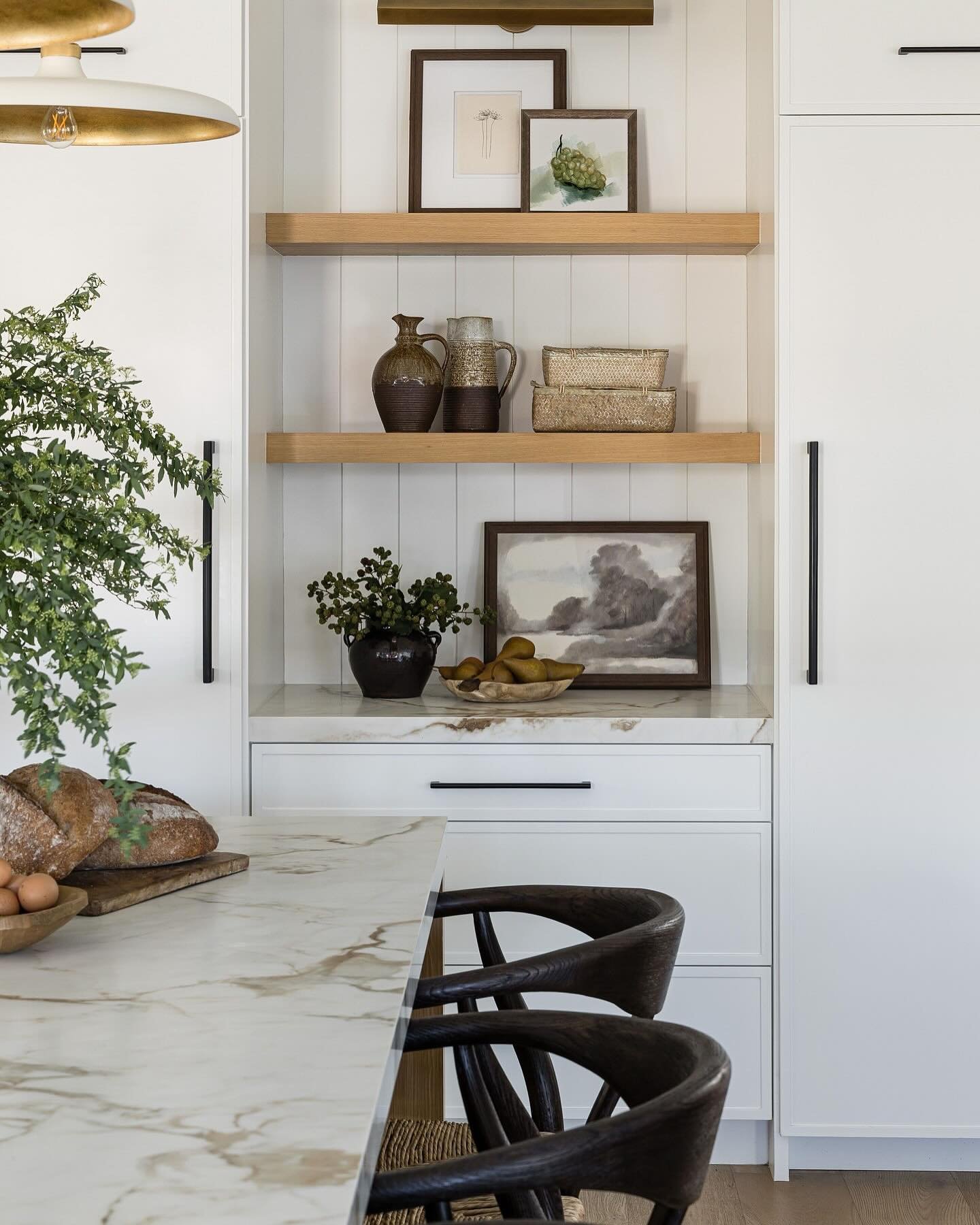 Two light wood floating shelves in a white kitchen nook with shiplap, styled with framed art and pottery.