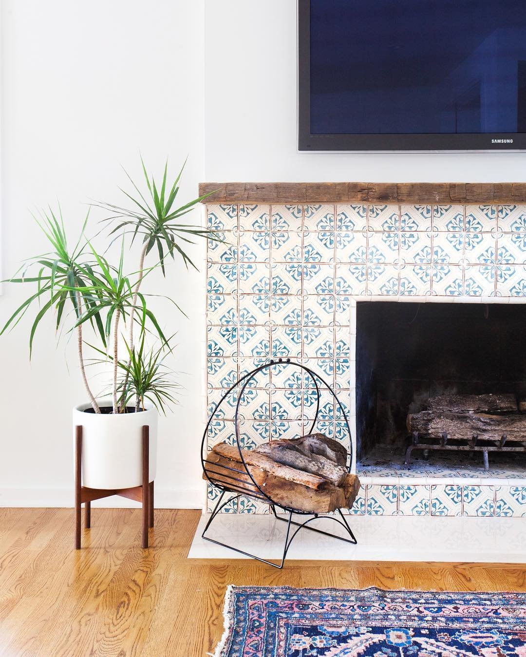 A contemporary fireplace with a rustic wood mantel, featuring a surround tiled with white and blue patterned tiles that have a distressed or antique finish.