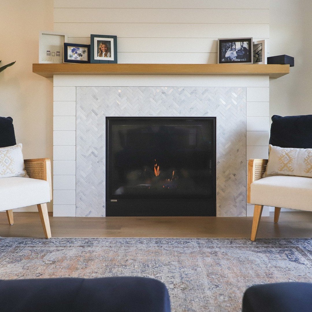 A modern fireplace with a light wood mantel and white shiplap trim, featuring a surround tiled with small white marble pieces in a herringbone pattern.