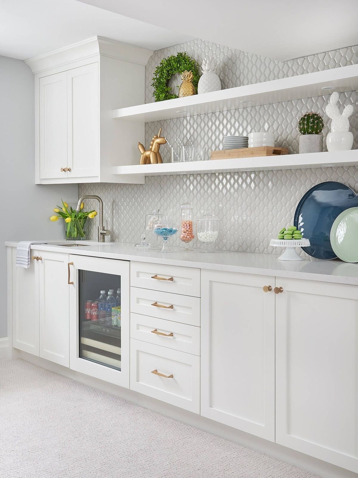 Two long white floating shelves mounted on a textured, reflective tile backsplash in a white kitchen bar.