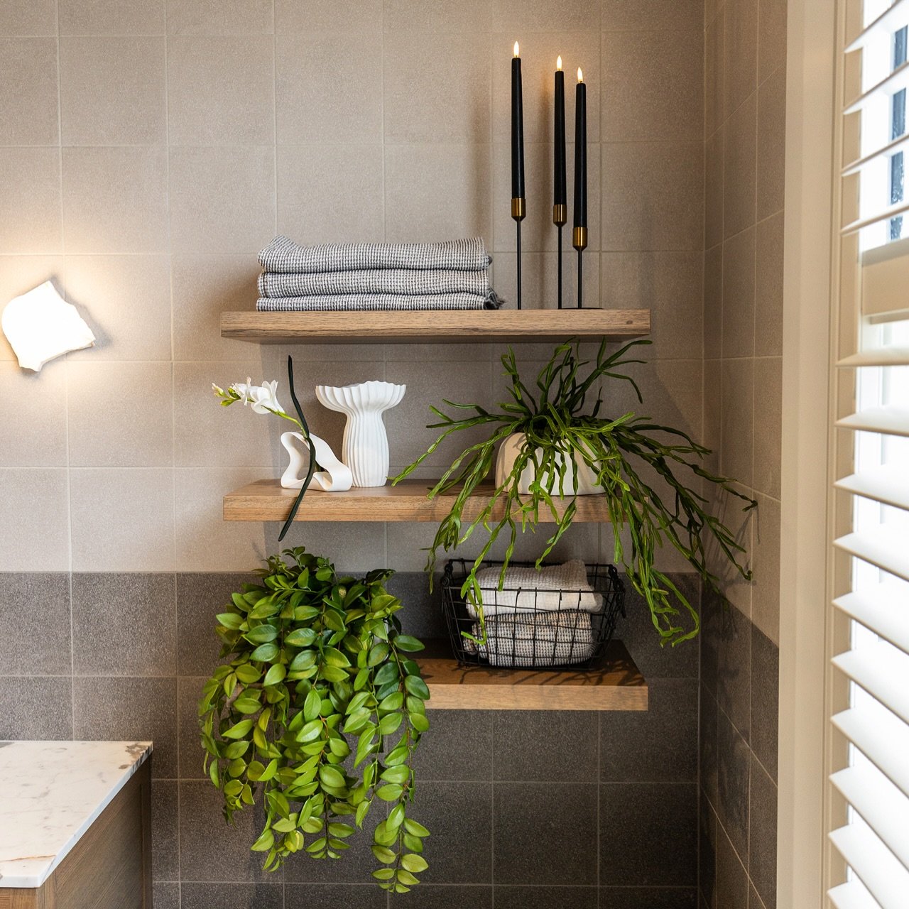 Three light wood floating shelves mounted on a two-tone grey tiled wall, styled with black candles, plants, and towels.