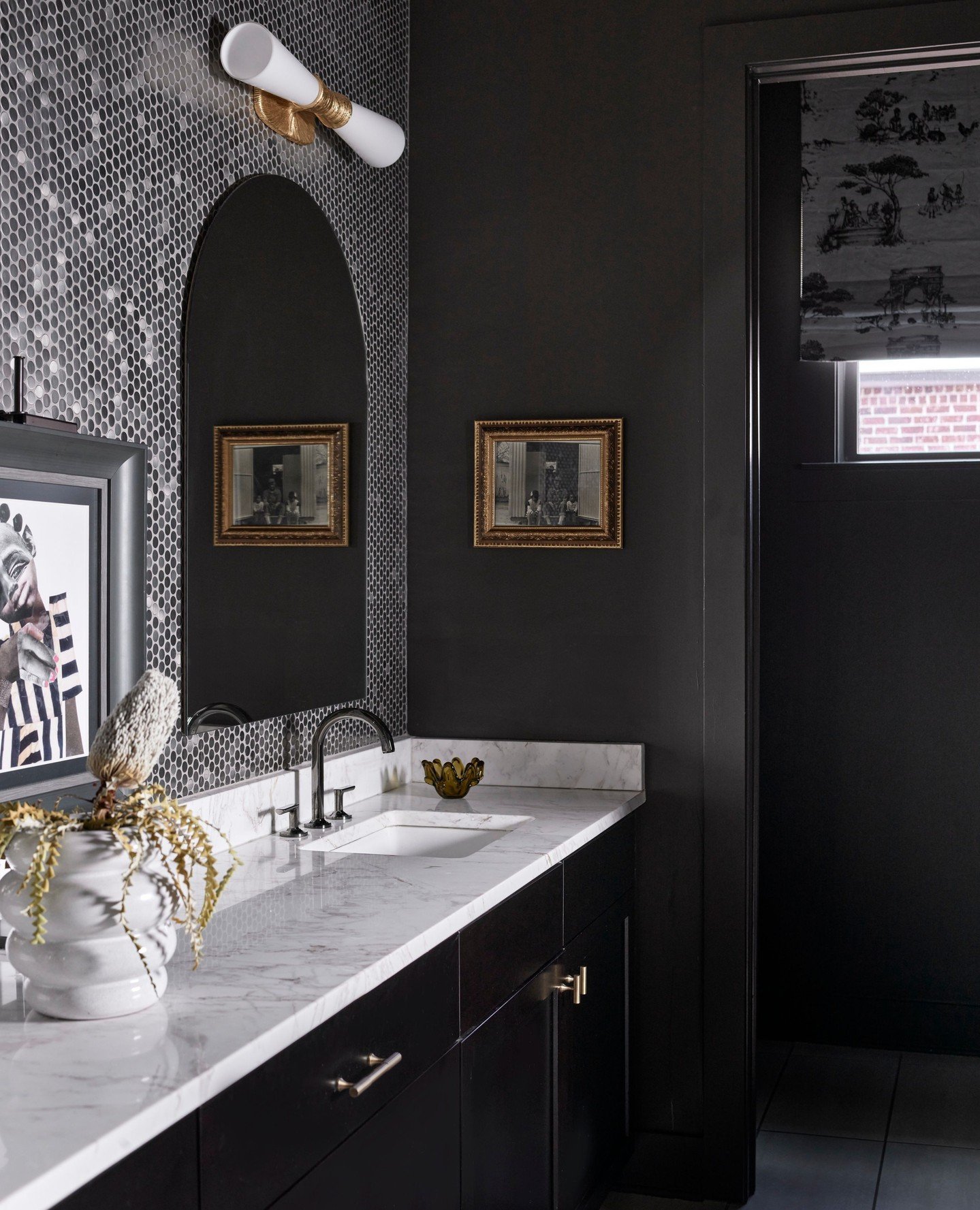 Horizontal white and brass barrel-shaped sconce mounted above a tall, arched black mirror in a dark gray and black bathroom with mosaic tile.