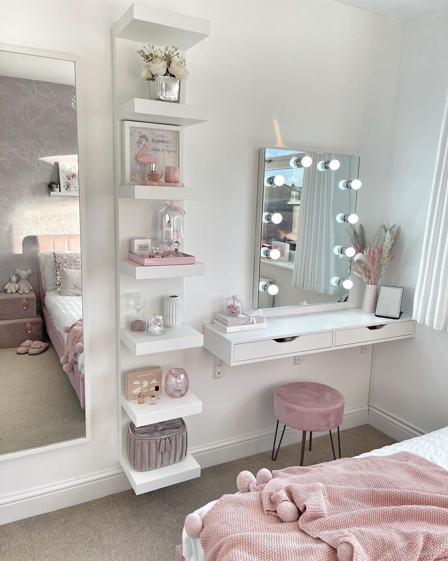 A compact white floating dressing table with a rectangular mirror featuring vanity lights, paired with vertical floating shelves for display and a small pink velvet stool.