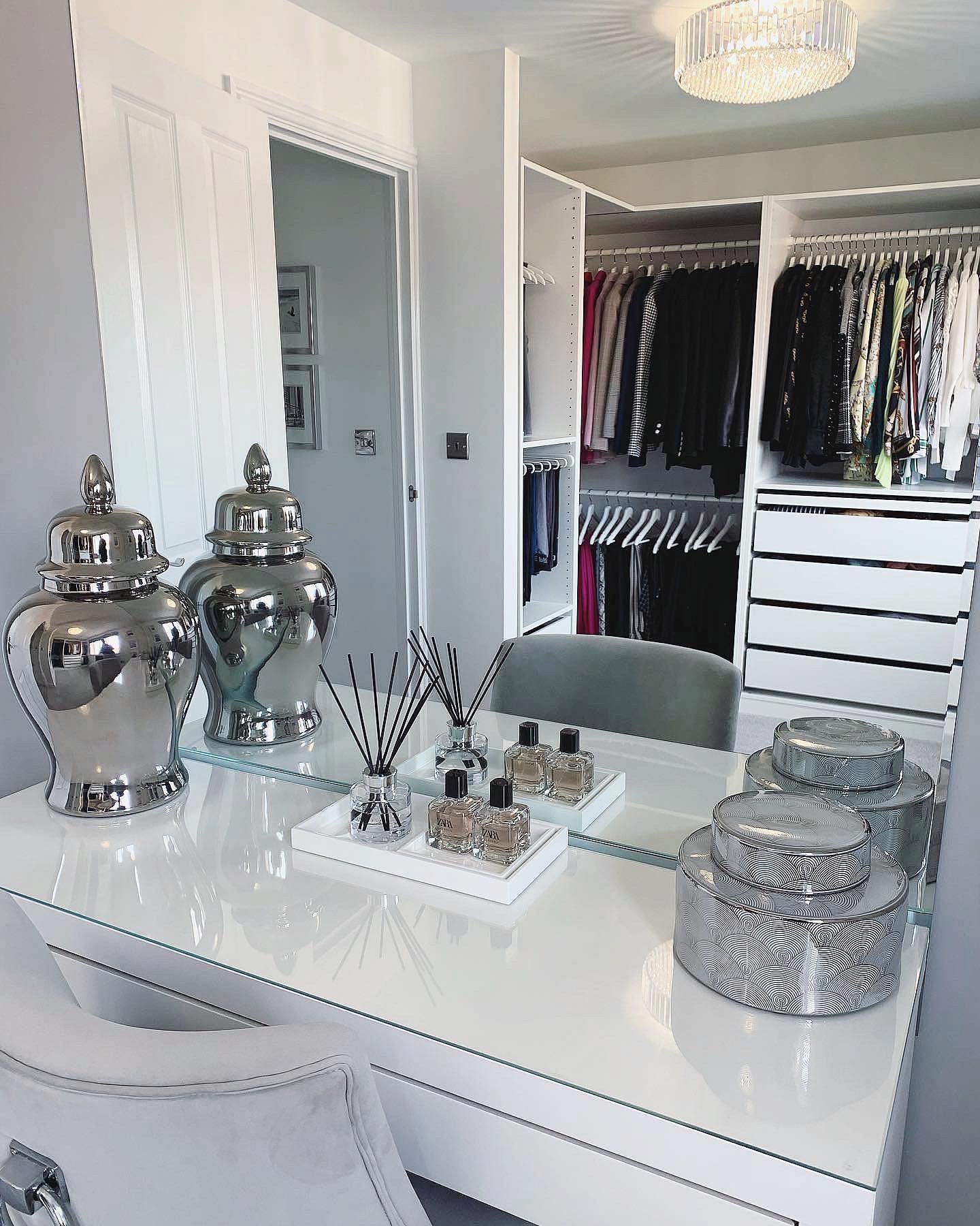 A white high-gloss vanity desk with silver decorative jars and perfumes, offering a direct view into an adjacent white walk-in closet.